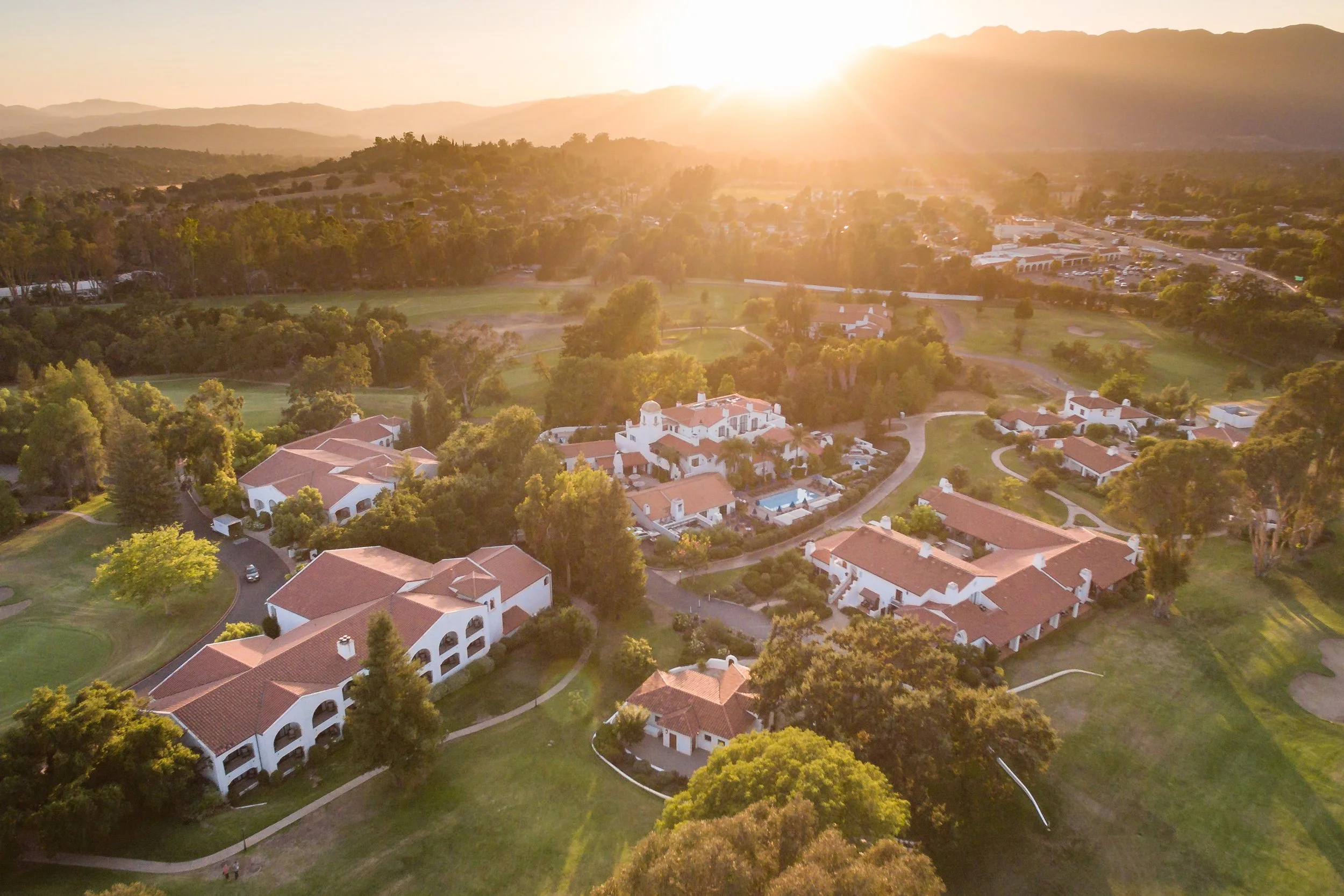 An aerial view of a suburban neighborhood at sunset with white houses and red-tiled roofs, surrounded by green trees and grassy areas, with mountains in the background.