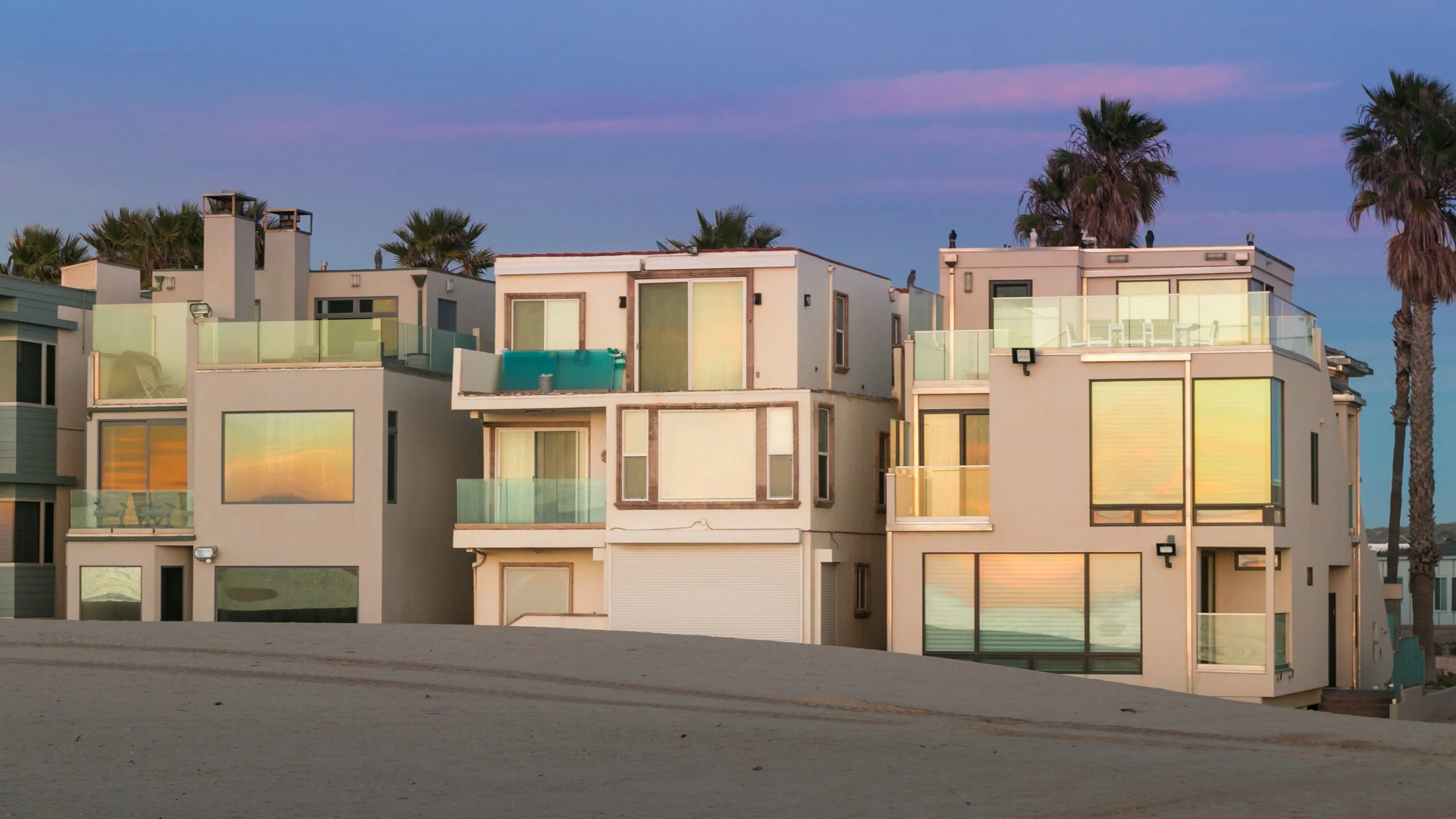 A row of modern multi-story apartment buildings with large glass windows and balconies, set against a sunset sky with palm trees in the background.