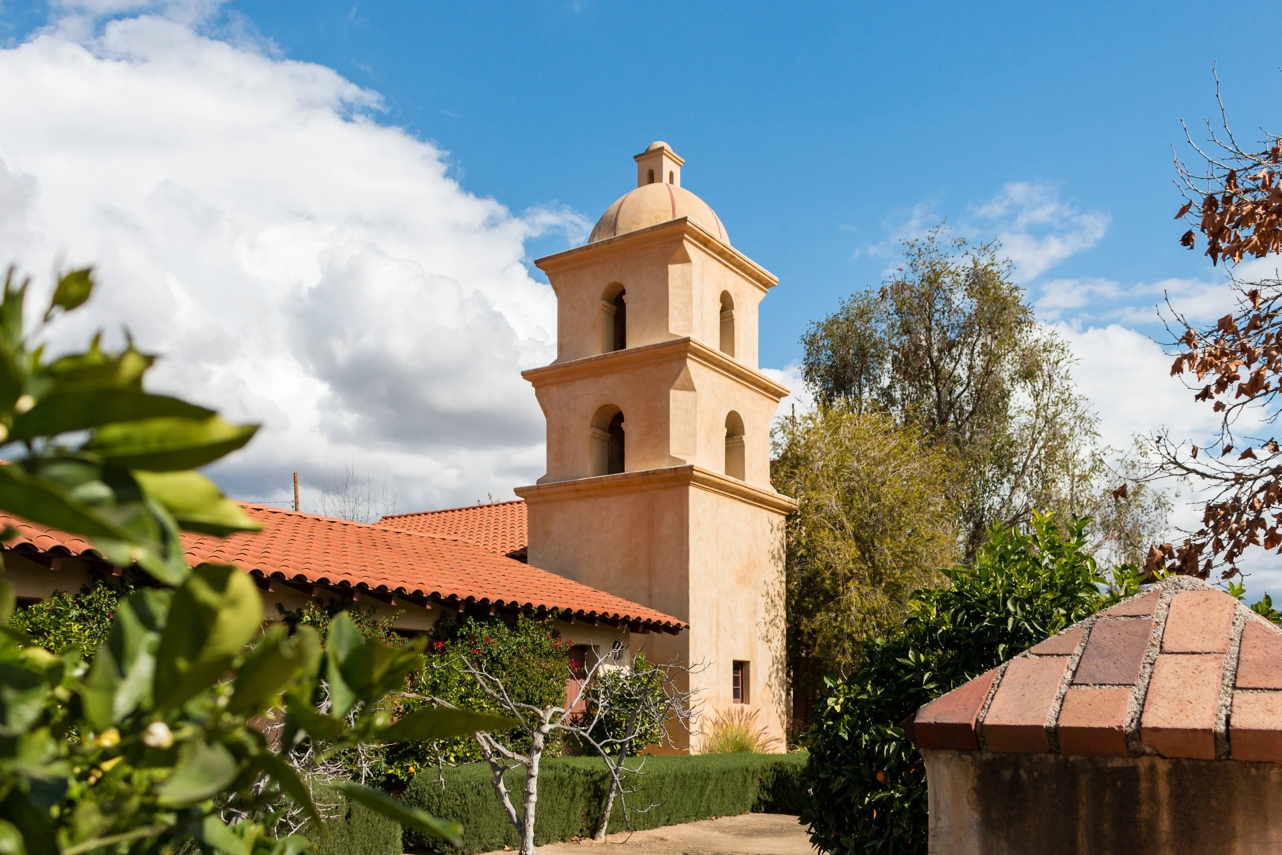 A beige stucco building with a tower featuring arched openings and a red tile roof, surrounded by green trees and plants, under a mostly cloudy sky.