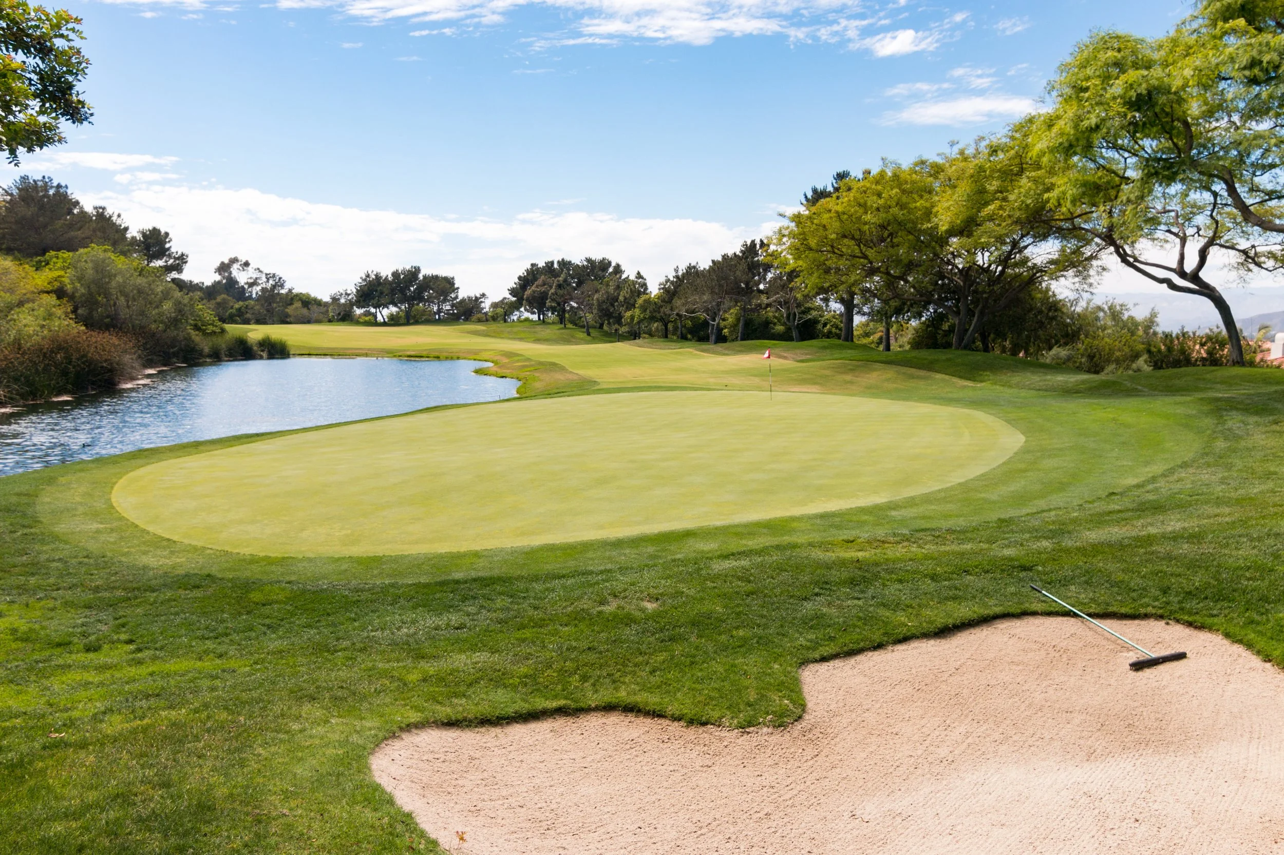 A golf course with a putting green, sand bunker, water hazard, and trees under a partly cloudy sky.