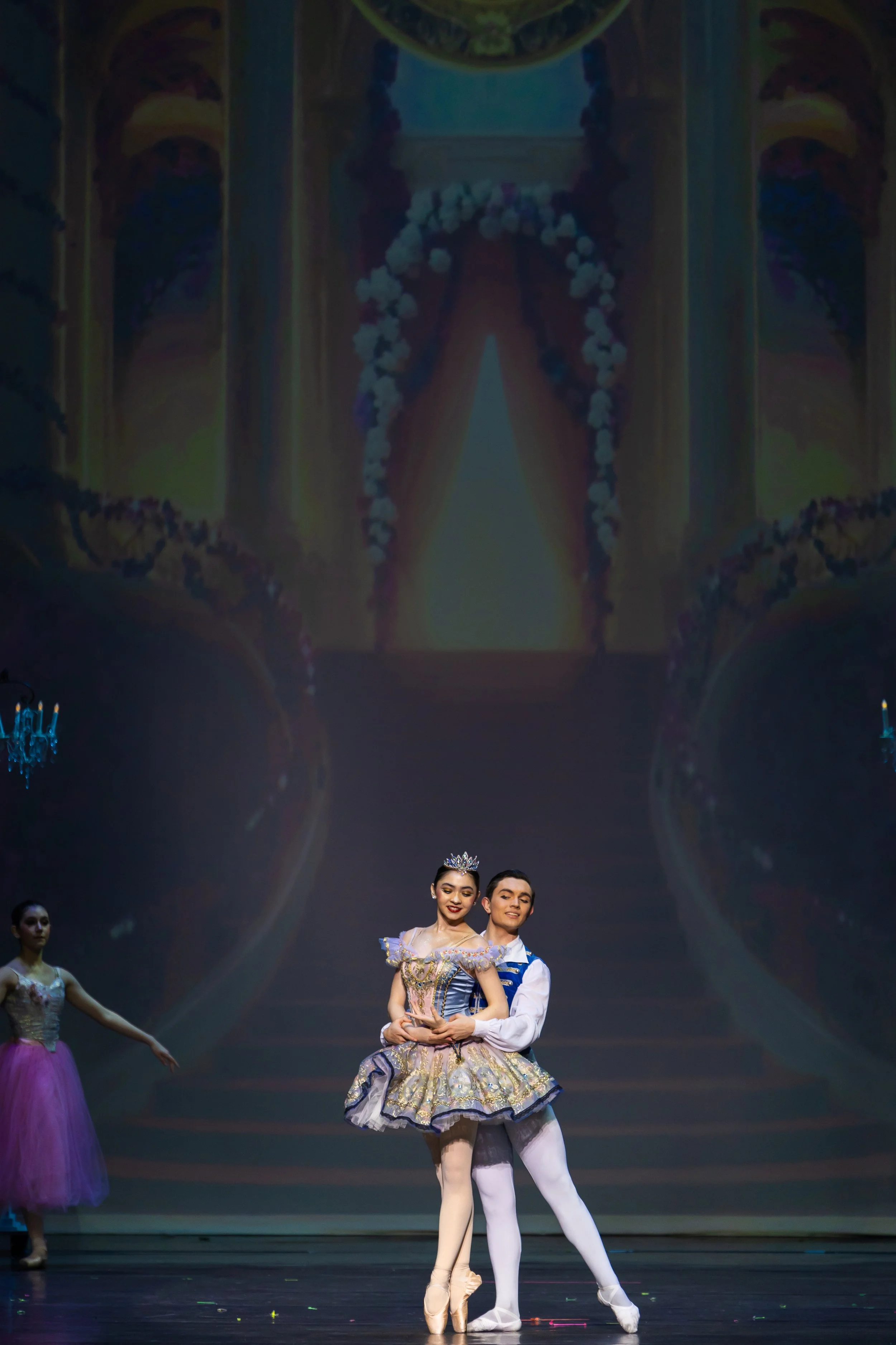 Ballet performance on stage, with a young woman and a young man in elegant costumes dancing together, a ballerina wearing a crown and tiara, stage decorated with large projection of a ballet scene.