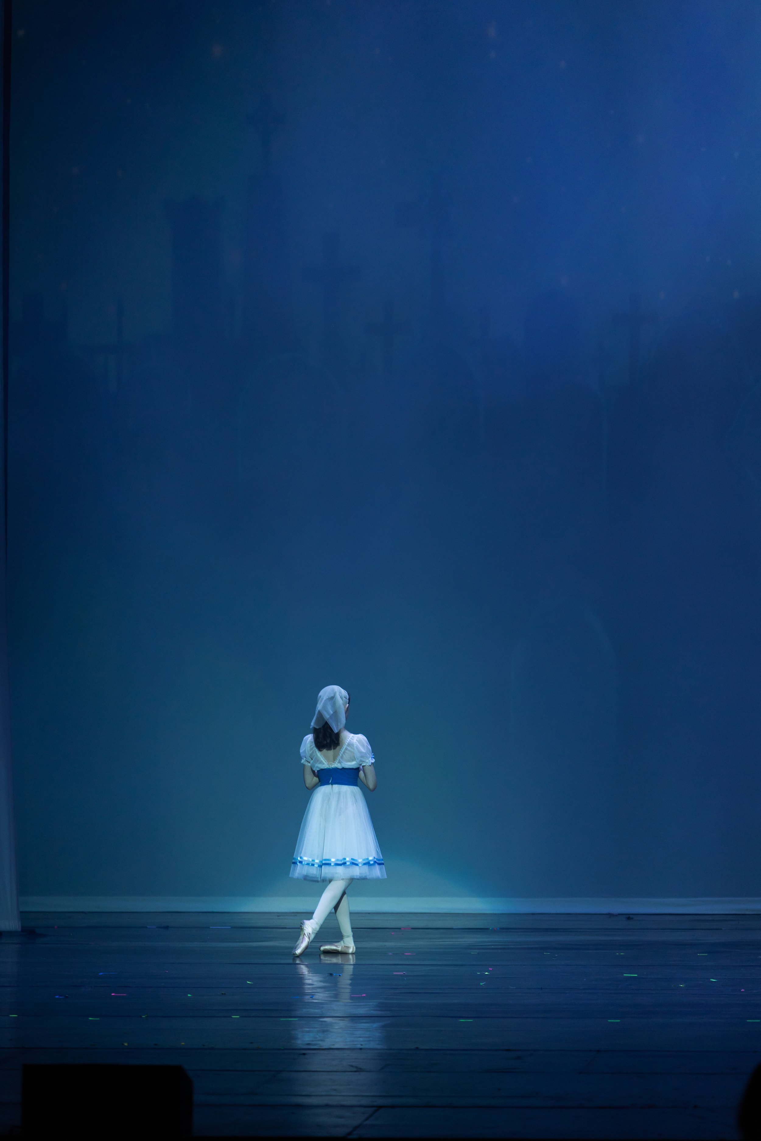 A ballerina in a white dress with blue accents performing on stage in front of a blue backdrop.