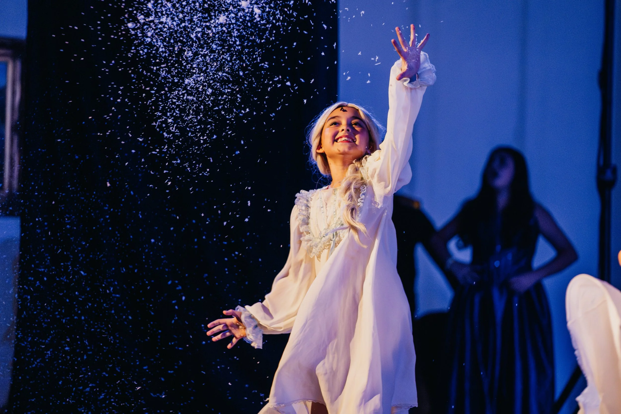 A woman with blonde hair in a white dress smiles and reaches her hand up as blue and white confetti falls around her in a dimly lit room.