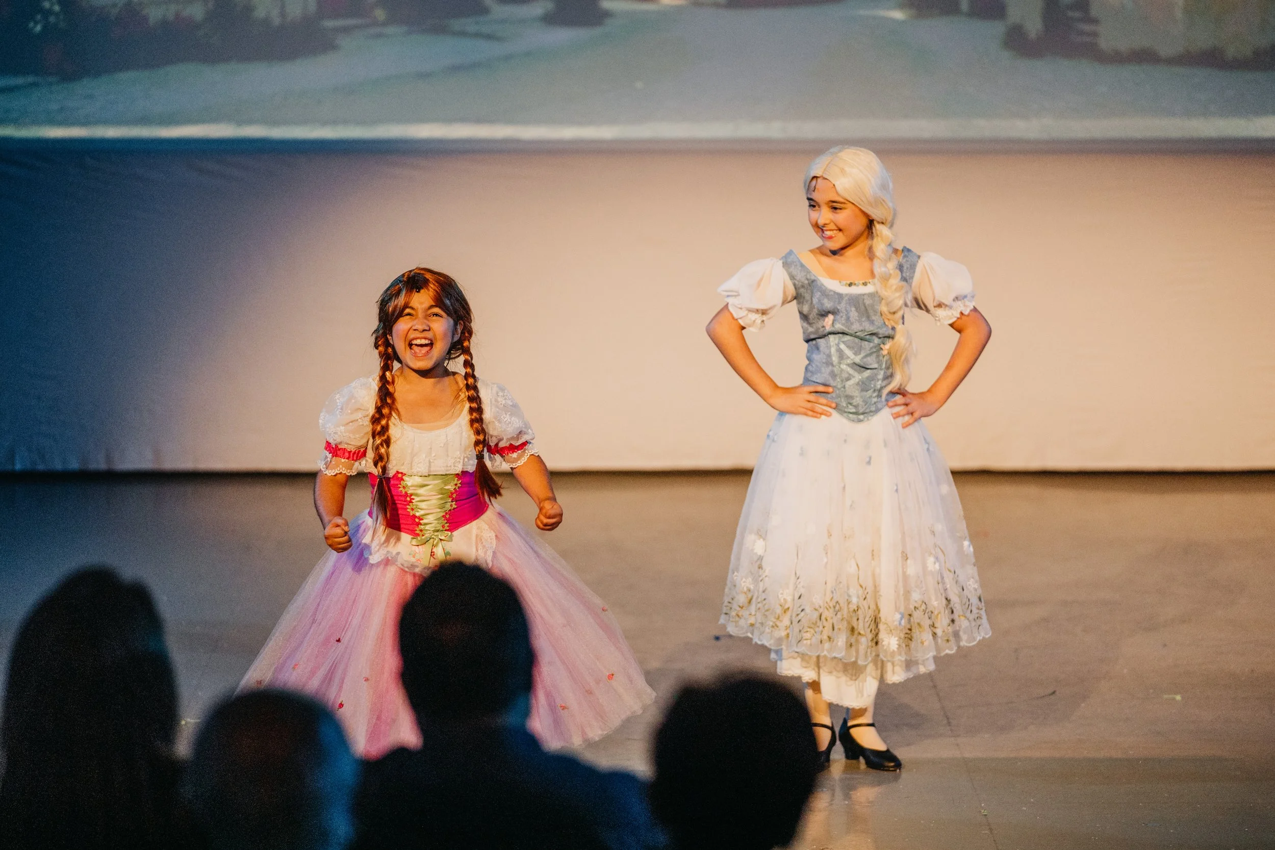 Two young girls dressed as princesses, one with long white hair and the other with braided hair, performing on stage during a play or costume event, with an audience in front.