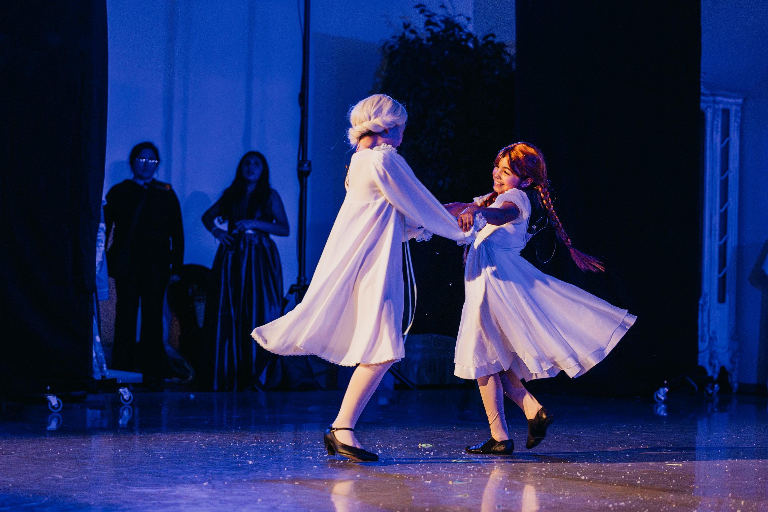 Two young girls dancing together on stage, wearing white dresses and black shoes, with stage lighting creating a dark blue ambience. Other people are observing in the background.
