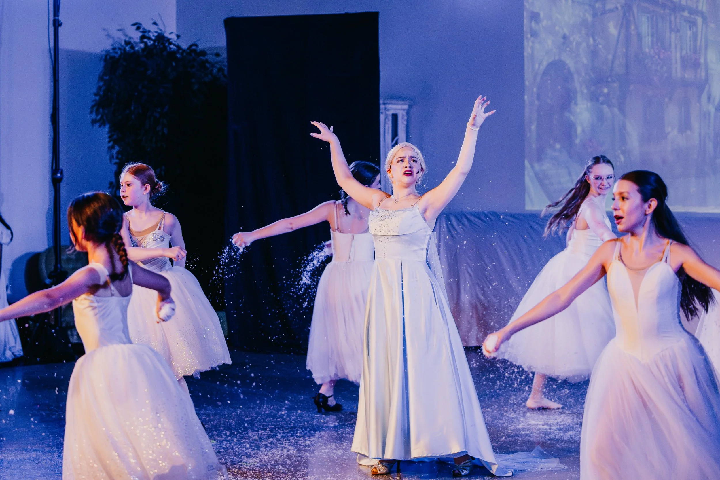 Ballet dancers performing on stage in white dresses, surrounded by water spray, with a blue background and stage set.