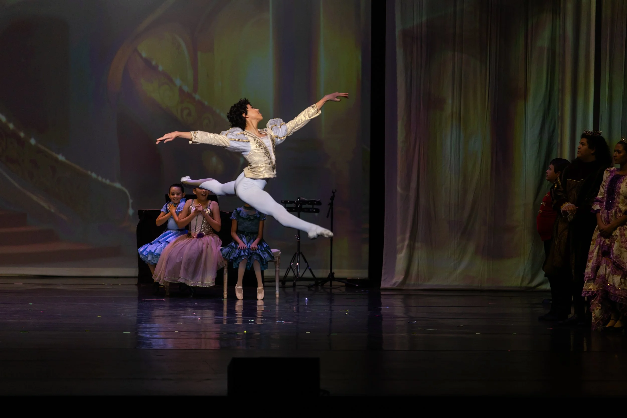Ballet dancer performing a leap on stage with a backdrop of a colorful projected scene, accompanied by children and adults in colorful costumes watching from the side.