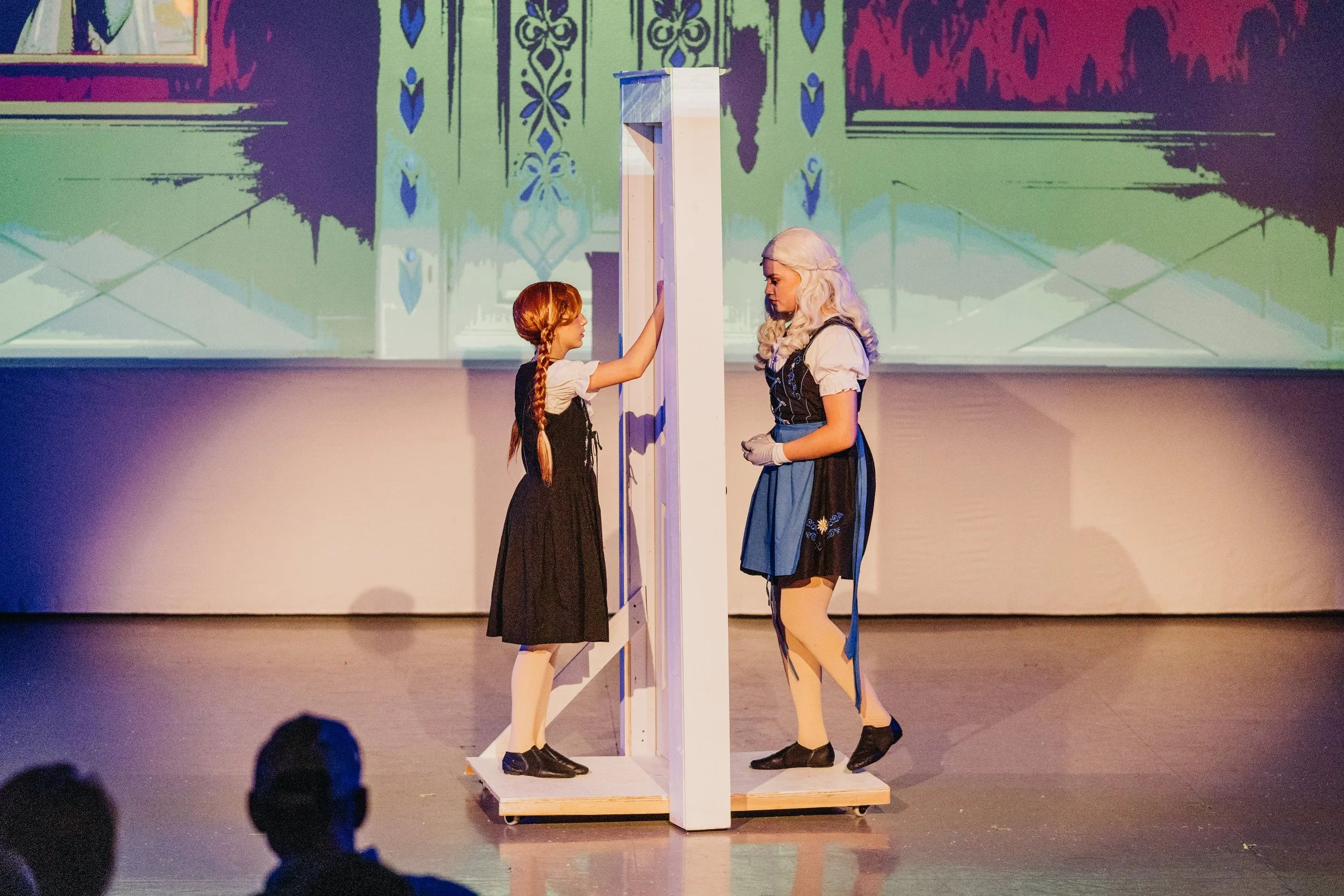 Two women dressed in traditional or historical clothing stand on a stage behind a wooden structure, possibly part of a theatrical or dance performance, with a background projected with colorful patterns.