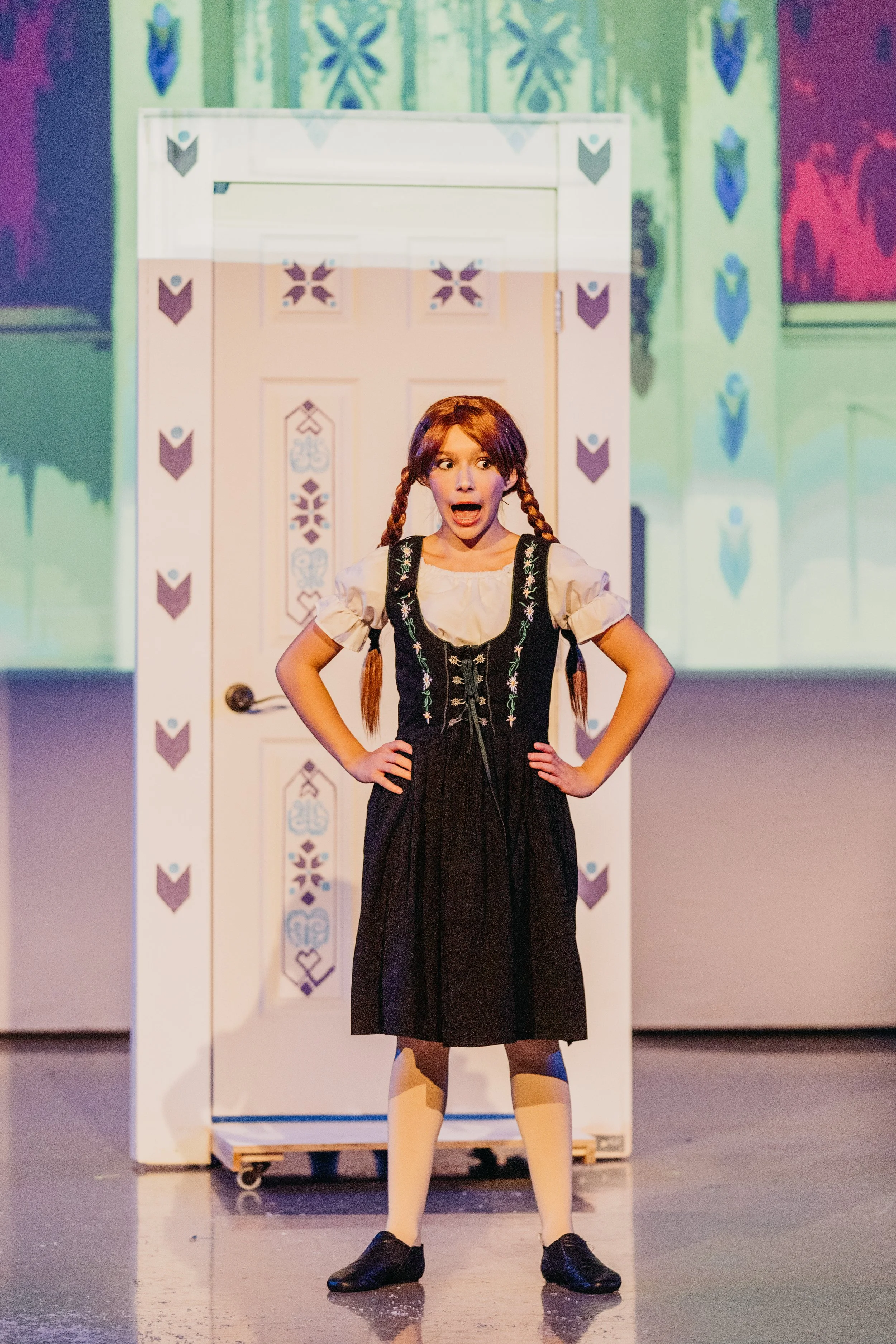 A young girl with red hair in braids, wearing a traditional dress with floral embroidery, white socks, and black shoes, stands with hands on hips in front of a decorated door during a theatrical performance.