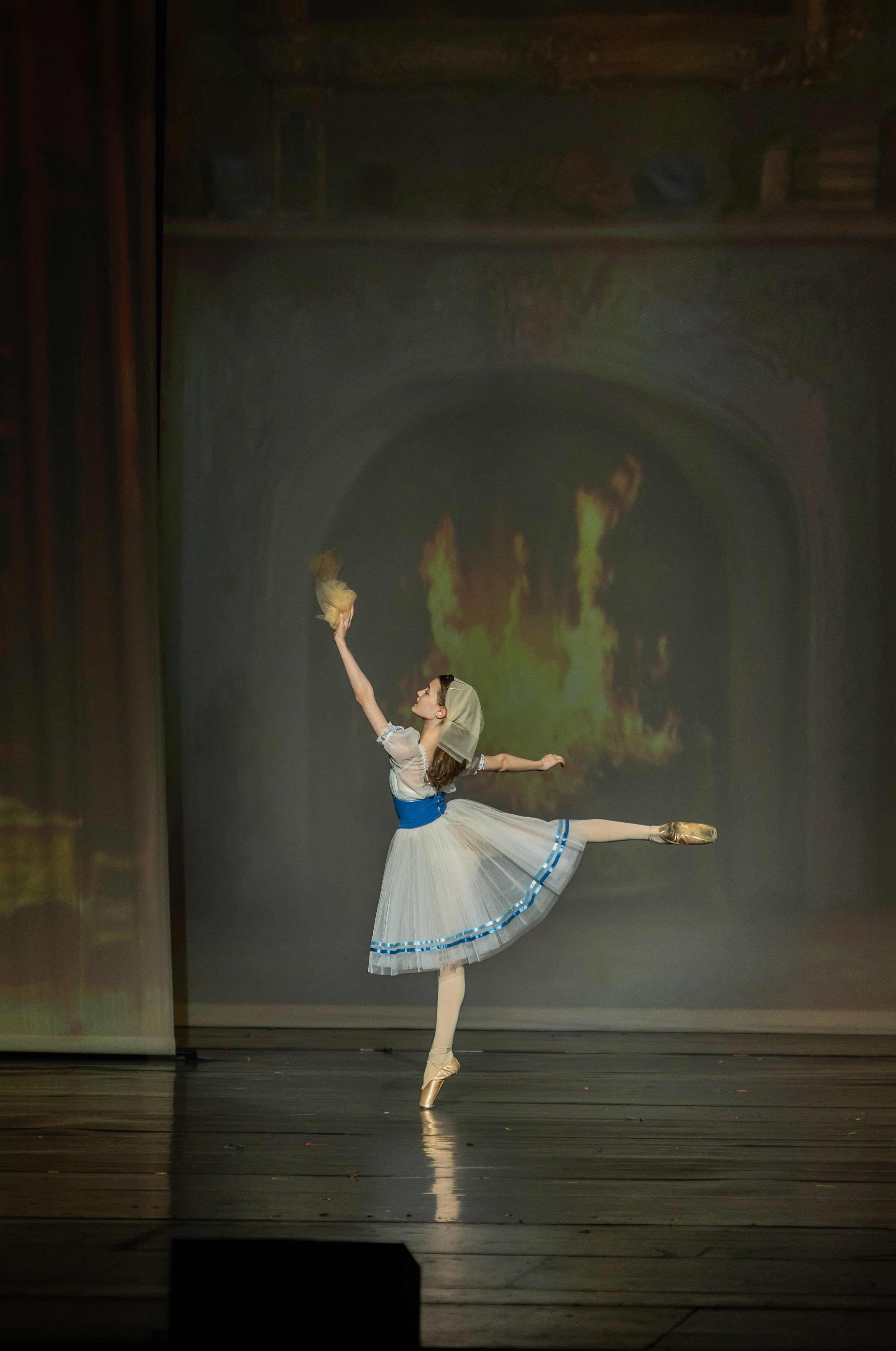 Ballet dancer performing on stage, wearing a white tutu with blue accents, balancing on one foot en pointe, in a classical ballet pose, with a backdrop displaying flames.