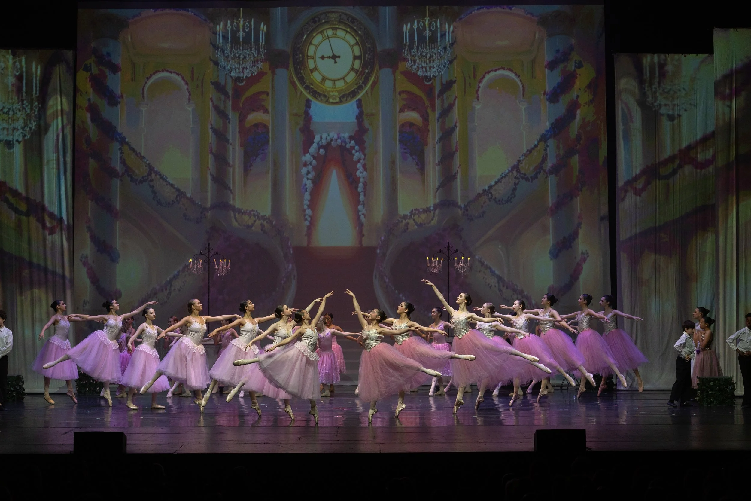 Ballet dancers in pink tutus performing on stage with a large backdrop of a grand ballroom with chandeliers, a clock, and ornate decorations.
