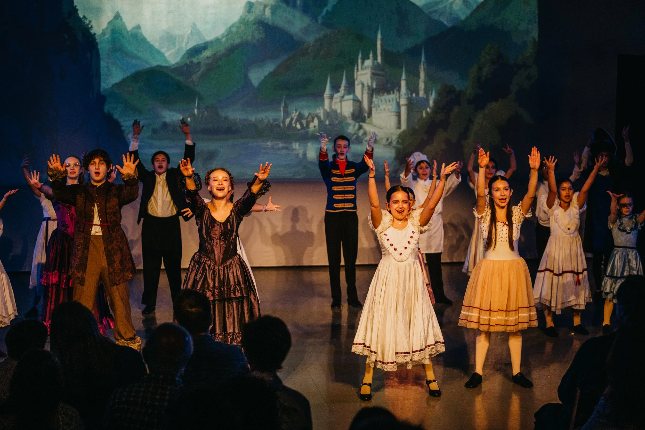 Children performing a play on stage in costumes from a historical or fairy tale era, with a scenic backdrop of a castle and mountains.