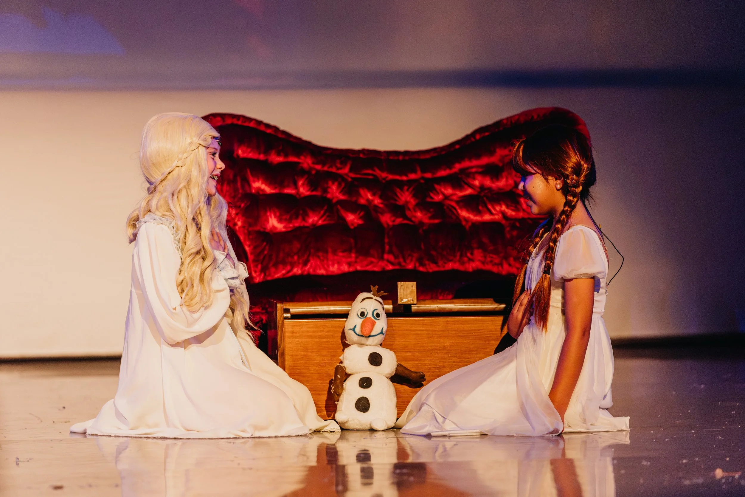 Two young girls dressed as princesses sit on the stage floor facing each other, holding hands. Between them is a snowman made of stuffed material with an orange nose, black eyes, and buttons. Behind them is a wooden box and a red velvet sofa, with a 