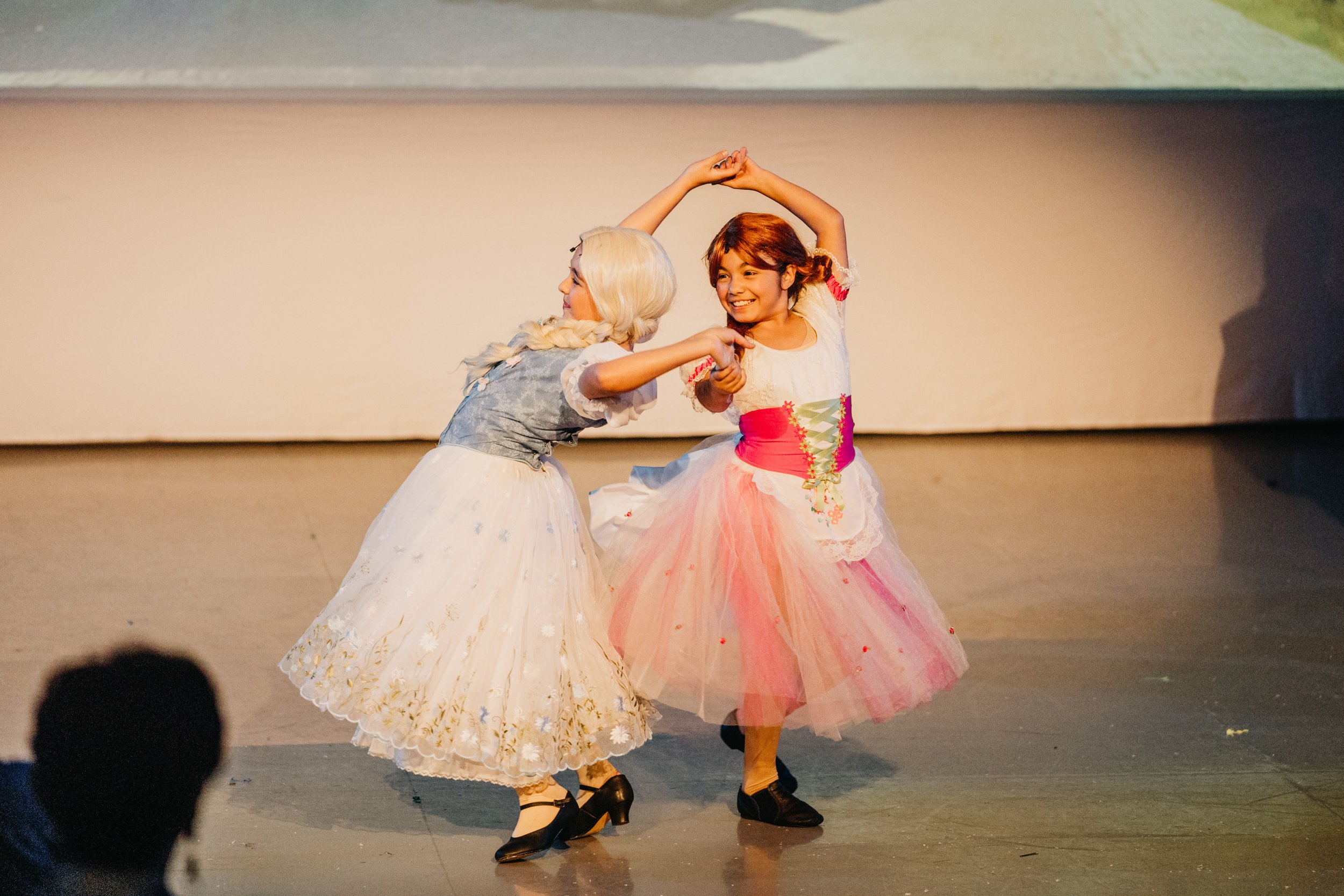 Two young girls in colorful princess costumes dance on stage, holding hands and smiling.