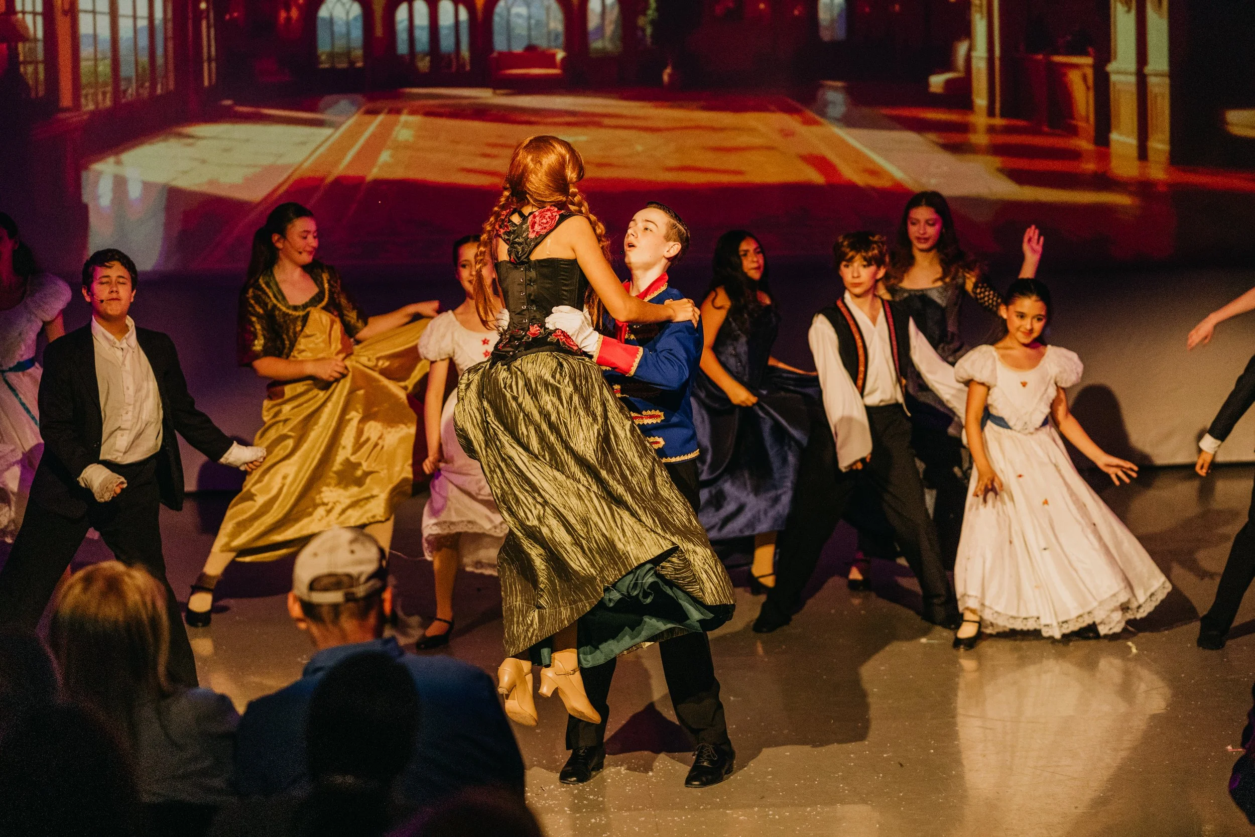 Performers dressed in historical costumes dancing on stage with a projected background of a grand hall.