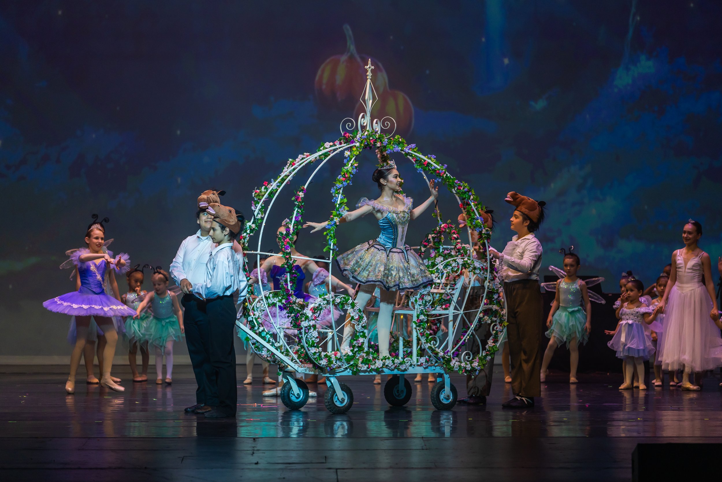 A stage with male and female performers dressed as characters from a fairy tale or ballet. The central figure is a woman in a colorful, ornate dress, standing in a decorative, flower-adorned carriage with large wheels. She is surrounded by children w