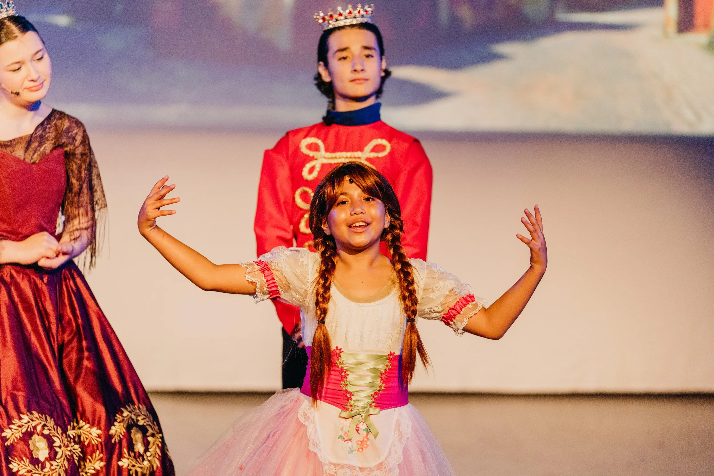 Young girl with long braided hair wearing a pink and white costume performing on stage with her arms raised, other performers in historical costumes are partially visible.