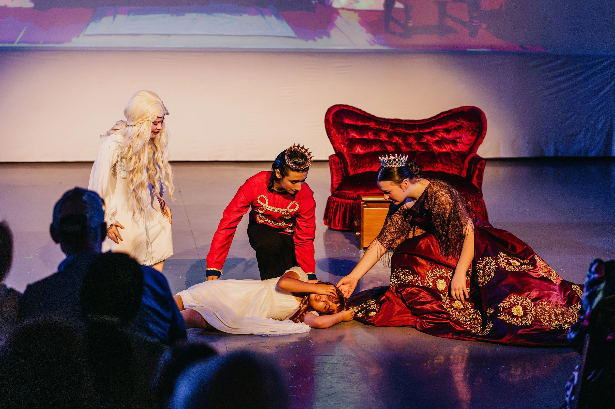 The image shows a theatrical scene with four women dressed in elaborate costumes. One woman is lying on the stage, seemingly unconscious or playing dead, while the other three are gathered around her, showing concern. The set includes a large, red, v