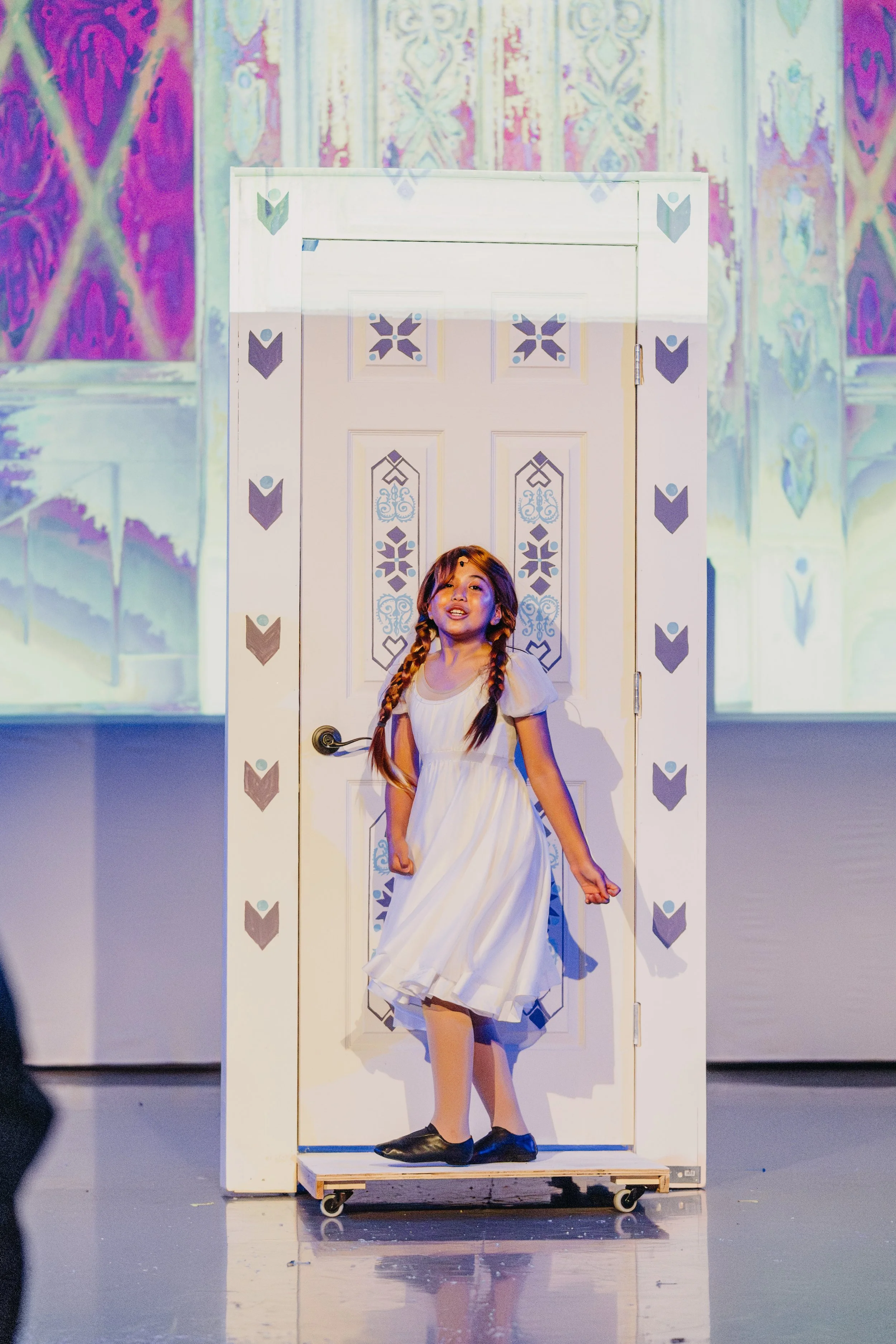 A young girl with brown hair in braids wearing a white dress, standing on a mobile platform in front of a decorated door with traditional patterns. She appears to be performing or posing.