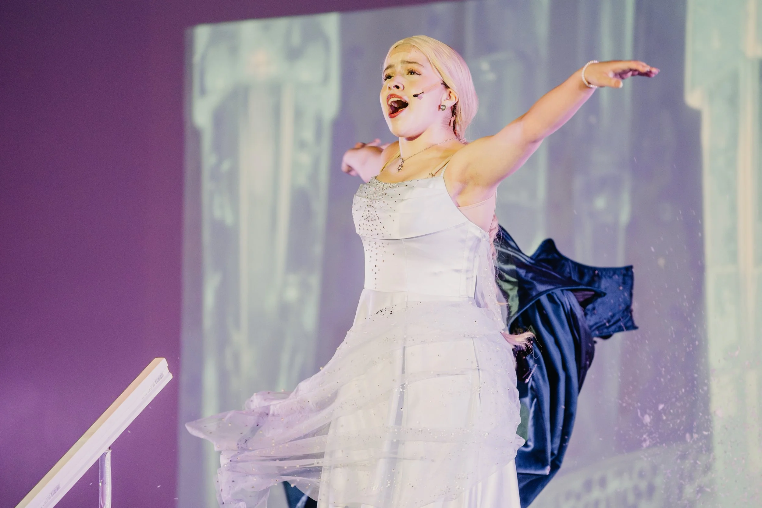 A performer with blonde hair wearing a white dress with rhinestones and a microphone headset singing with arms extended on stage.