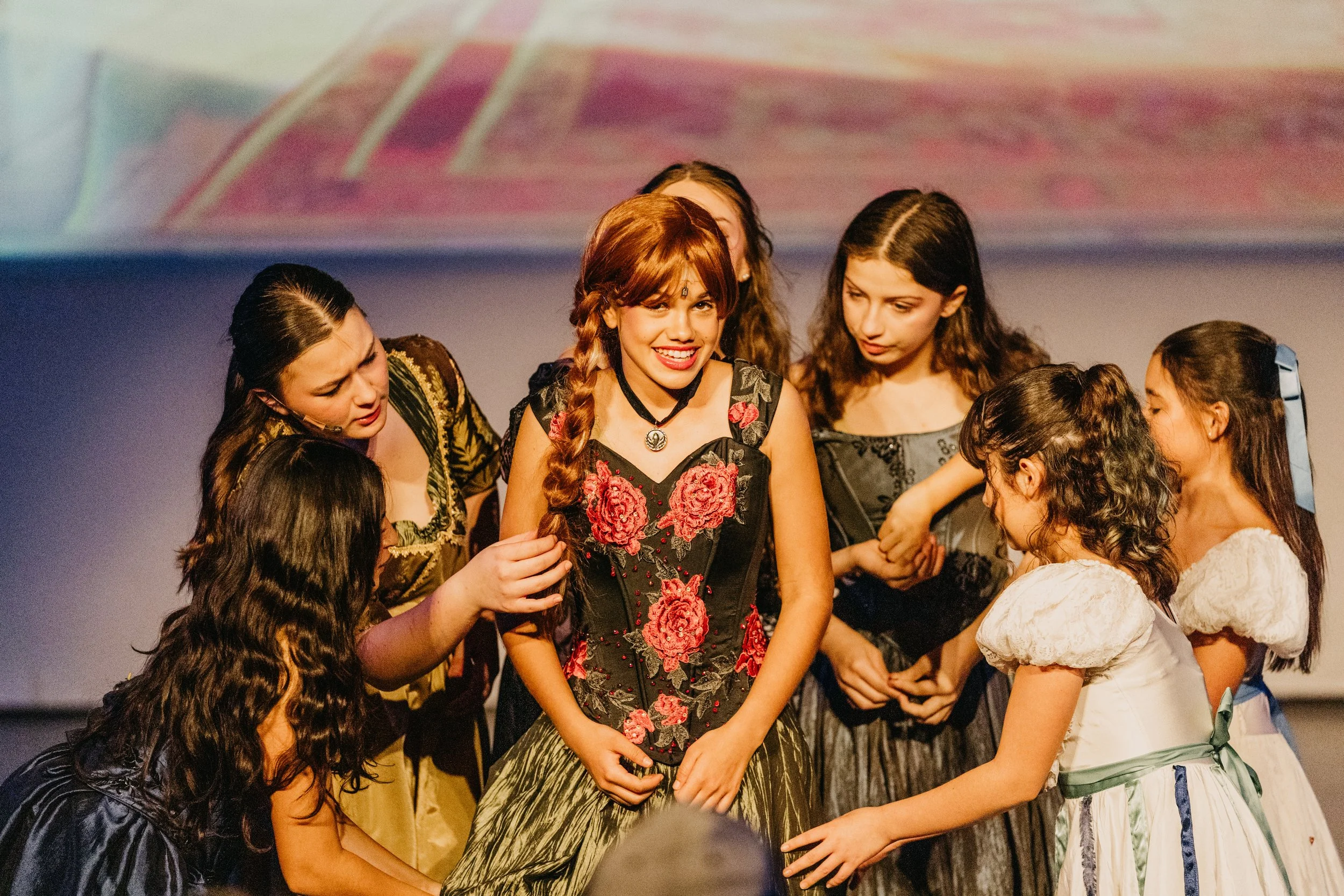 A group of young women wearing vintage-style dresses, gathered on a stage, with one woman in the center smiling and holding hands with others.
