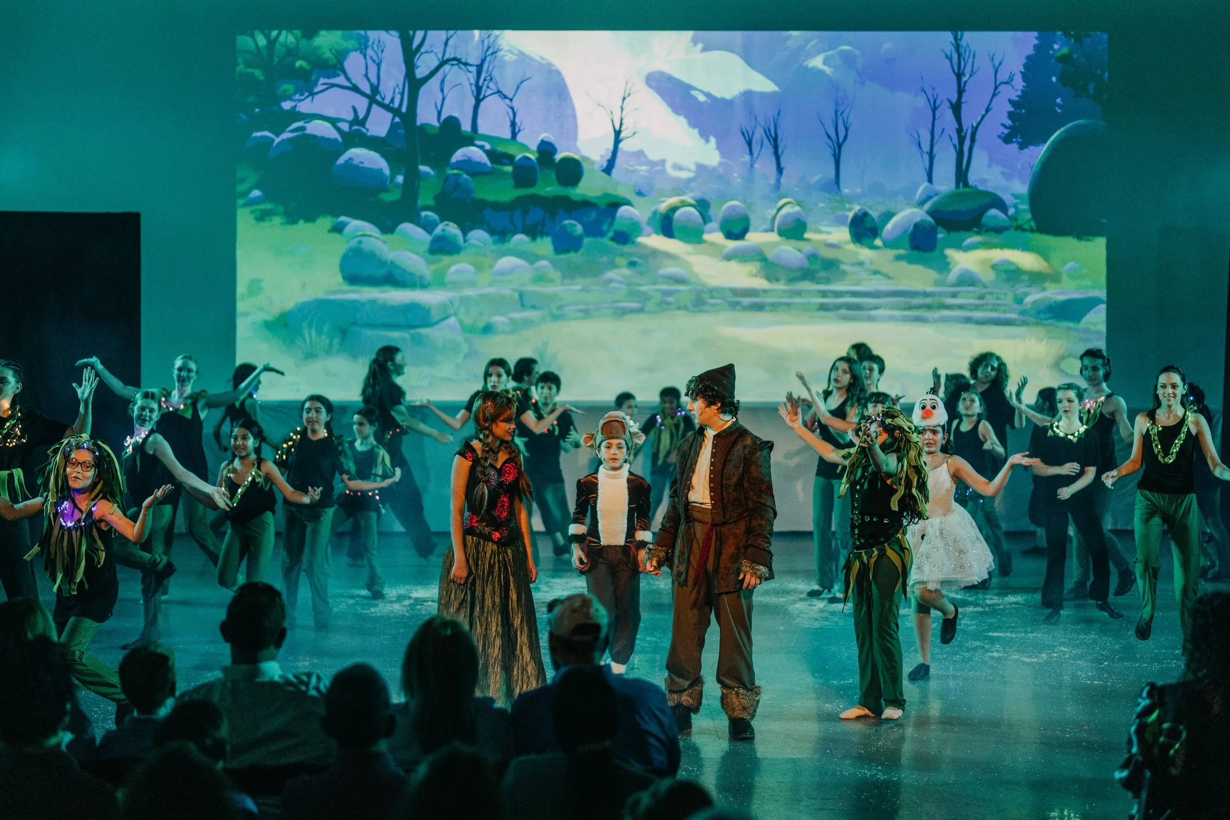 Children dressed in costumes performing a play or dance on stage with a digital backdrop of a scenic landscape with trees and rocks.