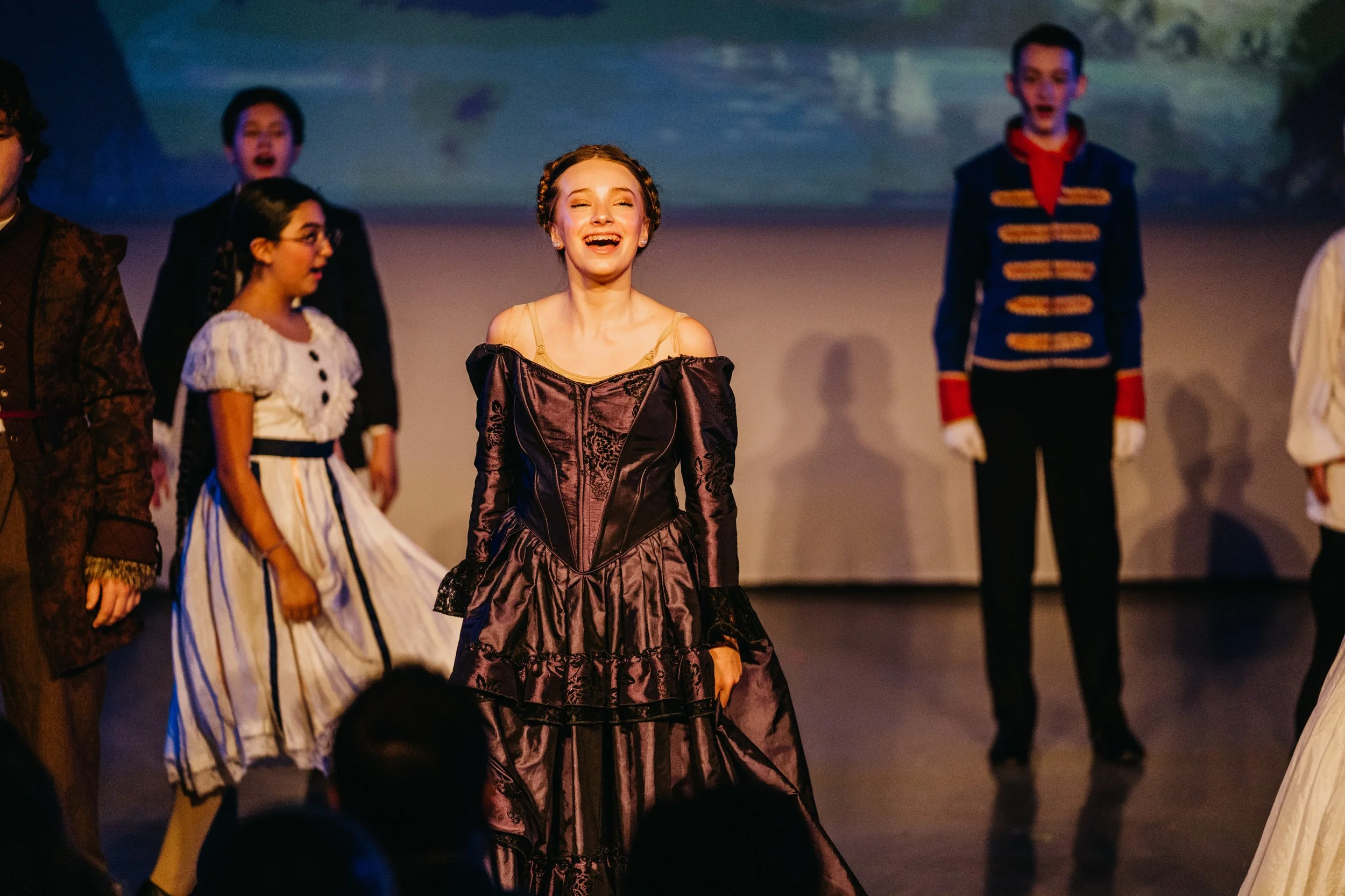 Actors in costume performing on stage, with a smiling woman in a dark Victorian-style dress in the foreground and other actors dressed in 19th-century attire in the background.