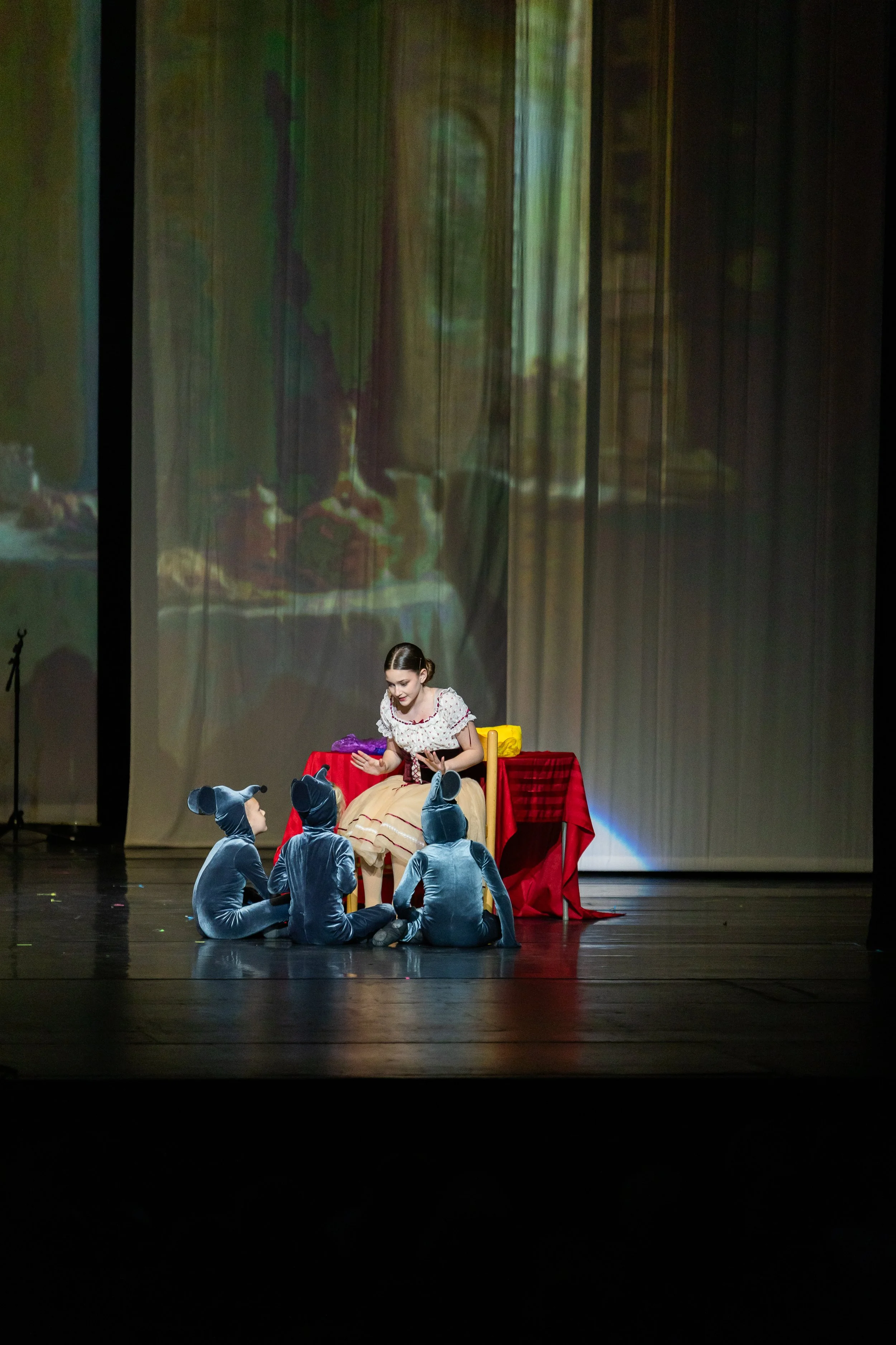 A theatrical performance on stage featuring a young actress dressed as Snow White sitting at a table with a red tablecloth, surrounded by four actors in mouse costumes. The backdrop displays a projected scene of a forest with greenery.