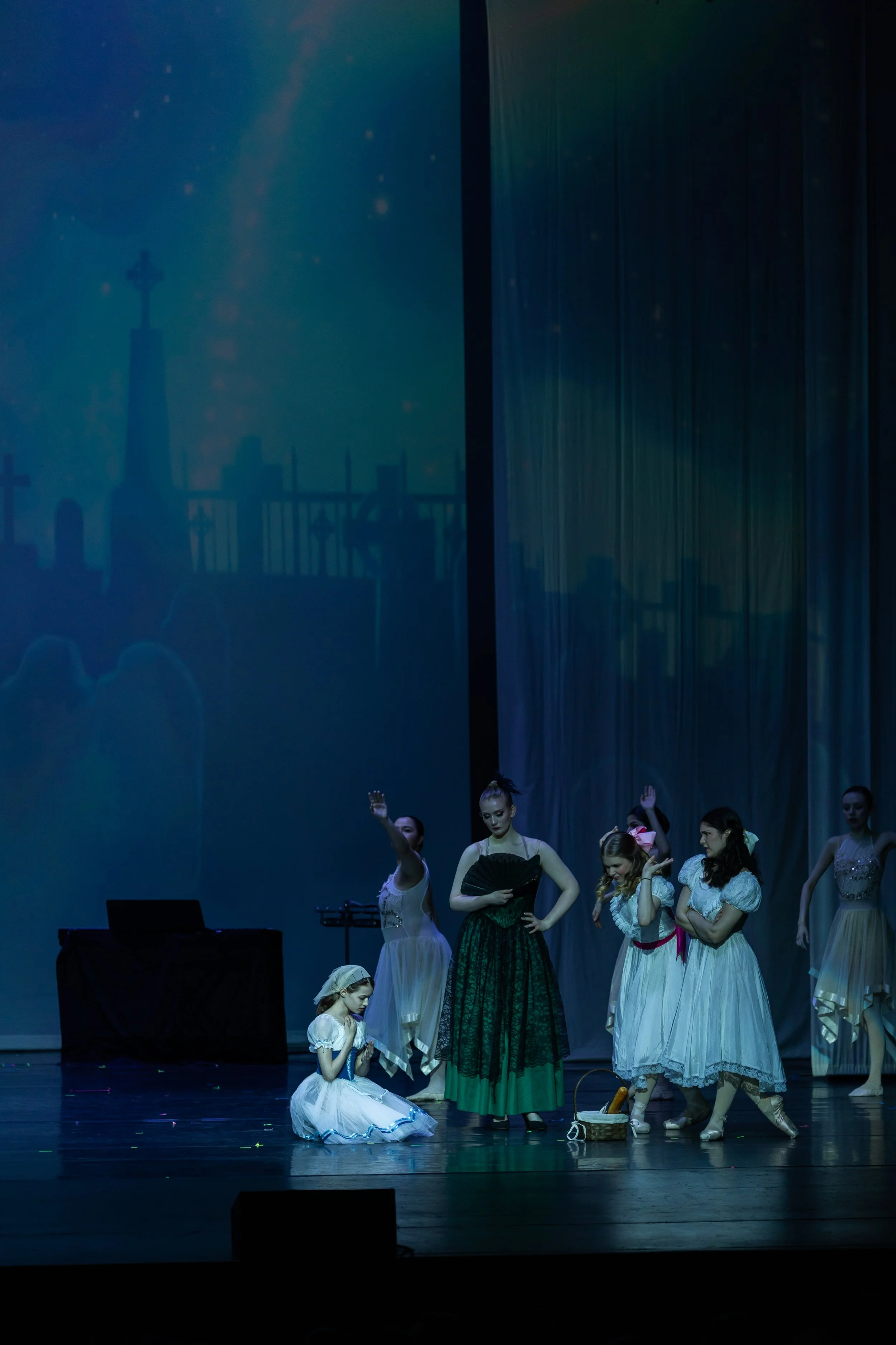Stage scene with ballet dancers in period costumes performing, with a backdrop displaying a church steeple and a cemetery.