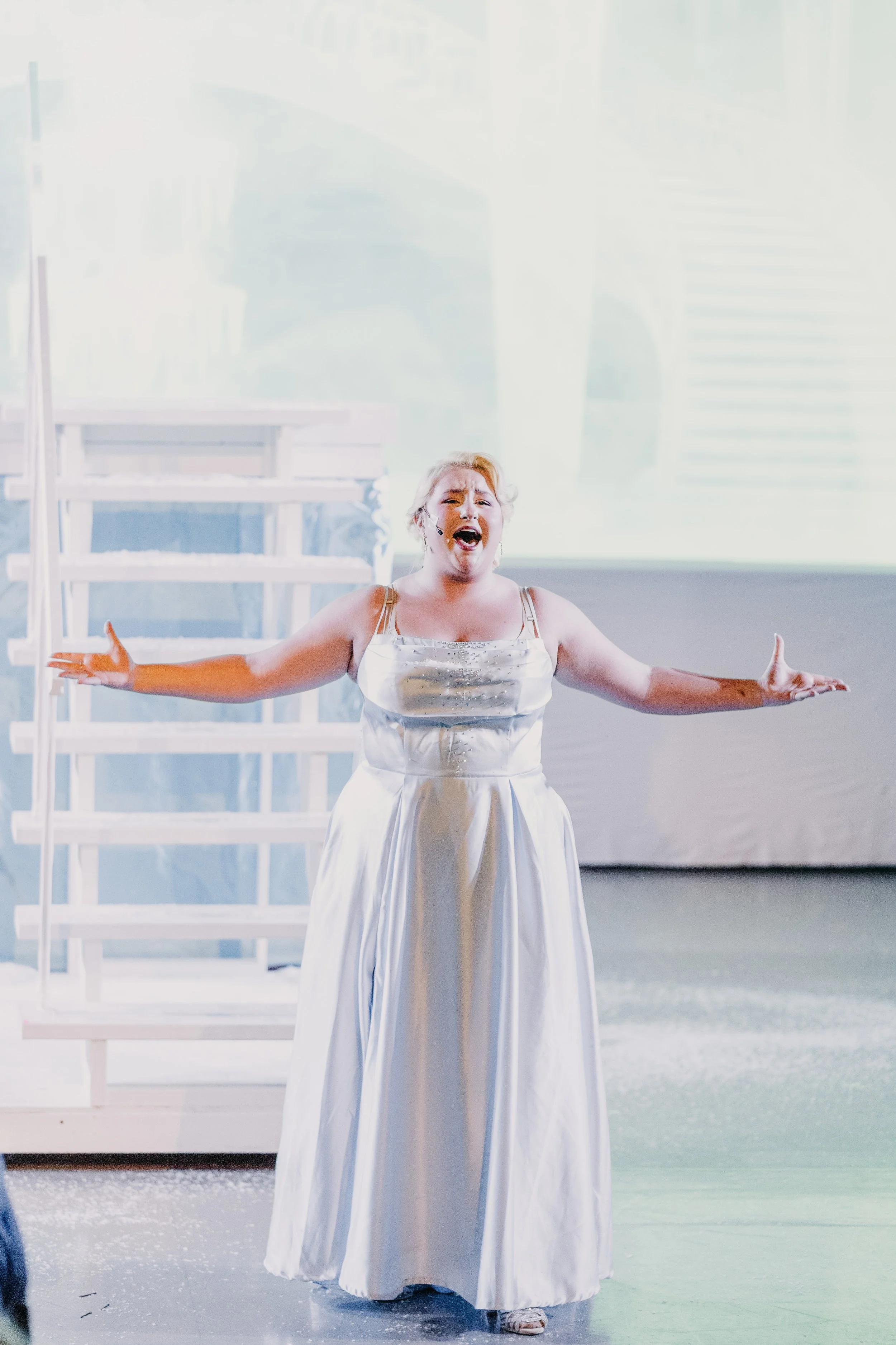 A woman wearing a white dress singing on stage with arms outstretched.