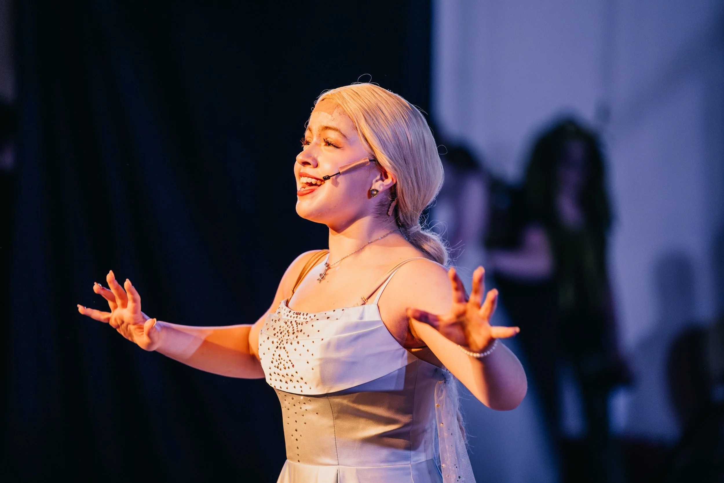 A woman with blonde hair in a braid, wearing a microphone headset, is performing with her arms extended outward and a joyful expression on her face in a stage setting.