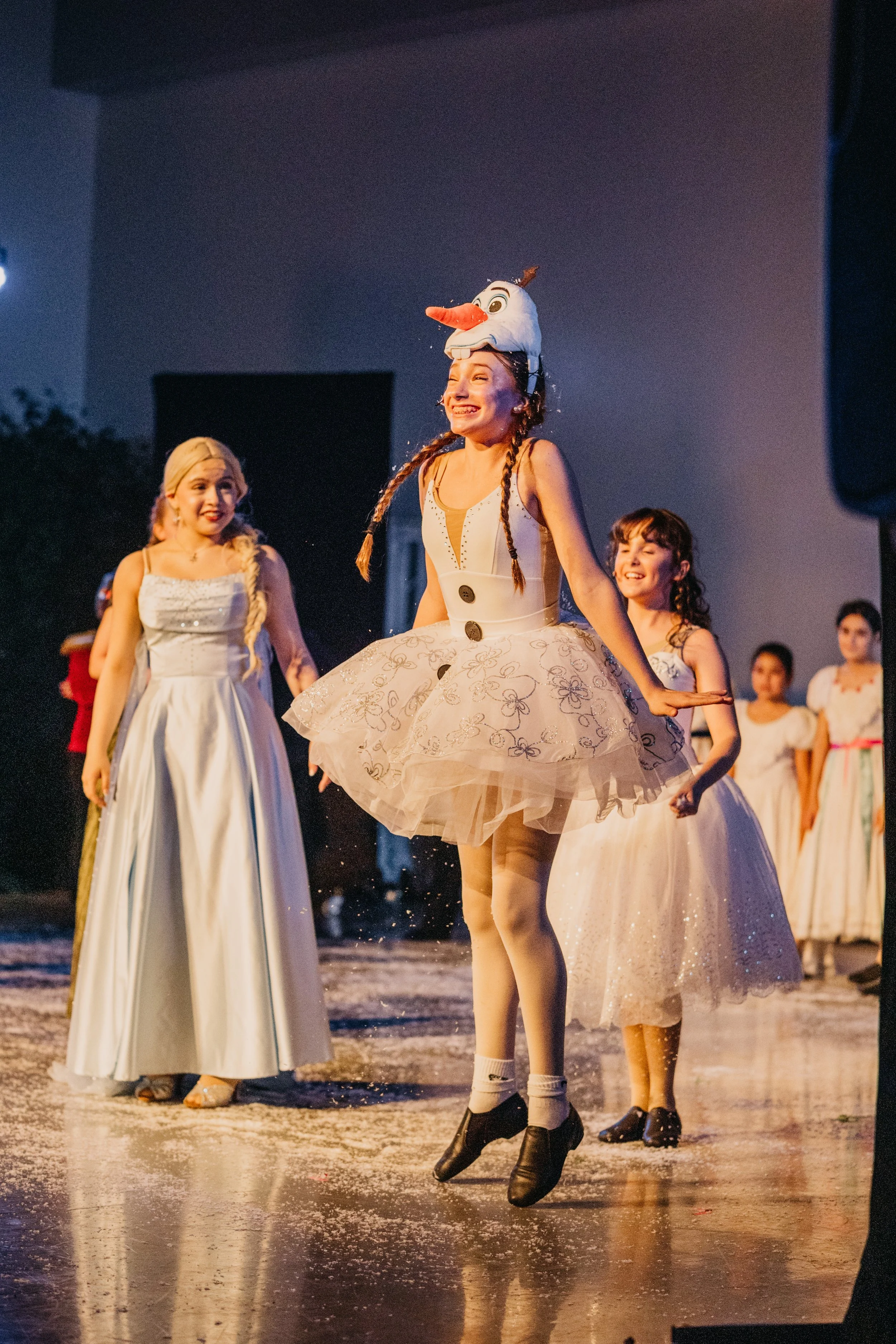 A young woman dressed as Olaf the snowman from Disney's Frozen, with a snowman face hat, is dancing among other girls in white dresses during a performance, with a joyful expression.