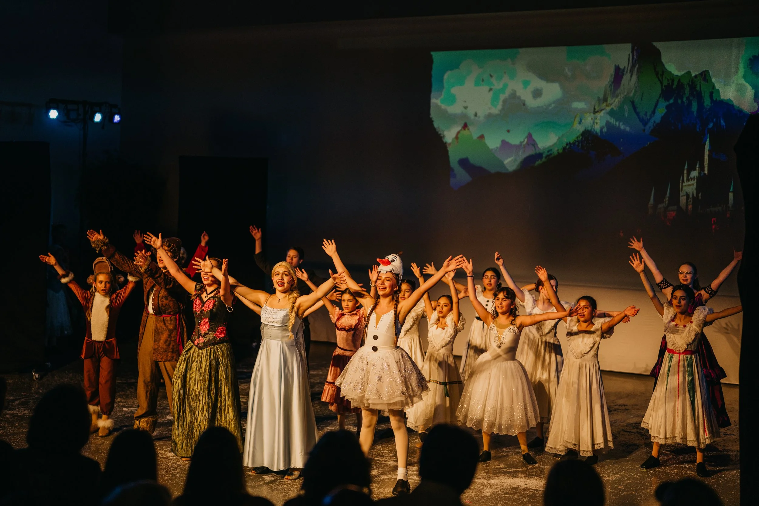 Group of children in costume performing on stage during a theatrical show, with a scenic mountain backdrop projected on the screen behind them.