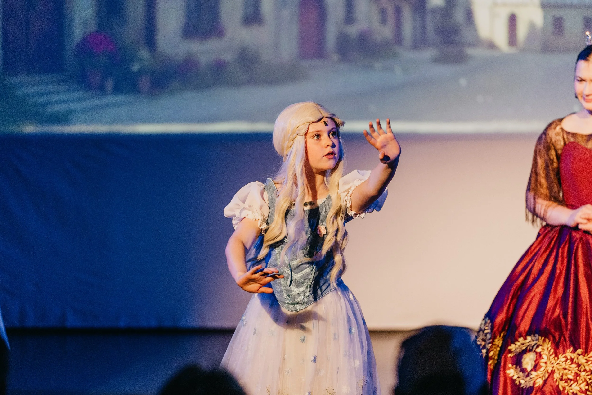 A young woman dressed as a princess with long blonde hair and a crown. She is wearing a white gown with floral embroidery and is gesturing with her right hand. Another woman dressed in a colorful, patterned costume is partially visible on the right s