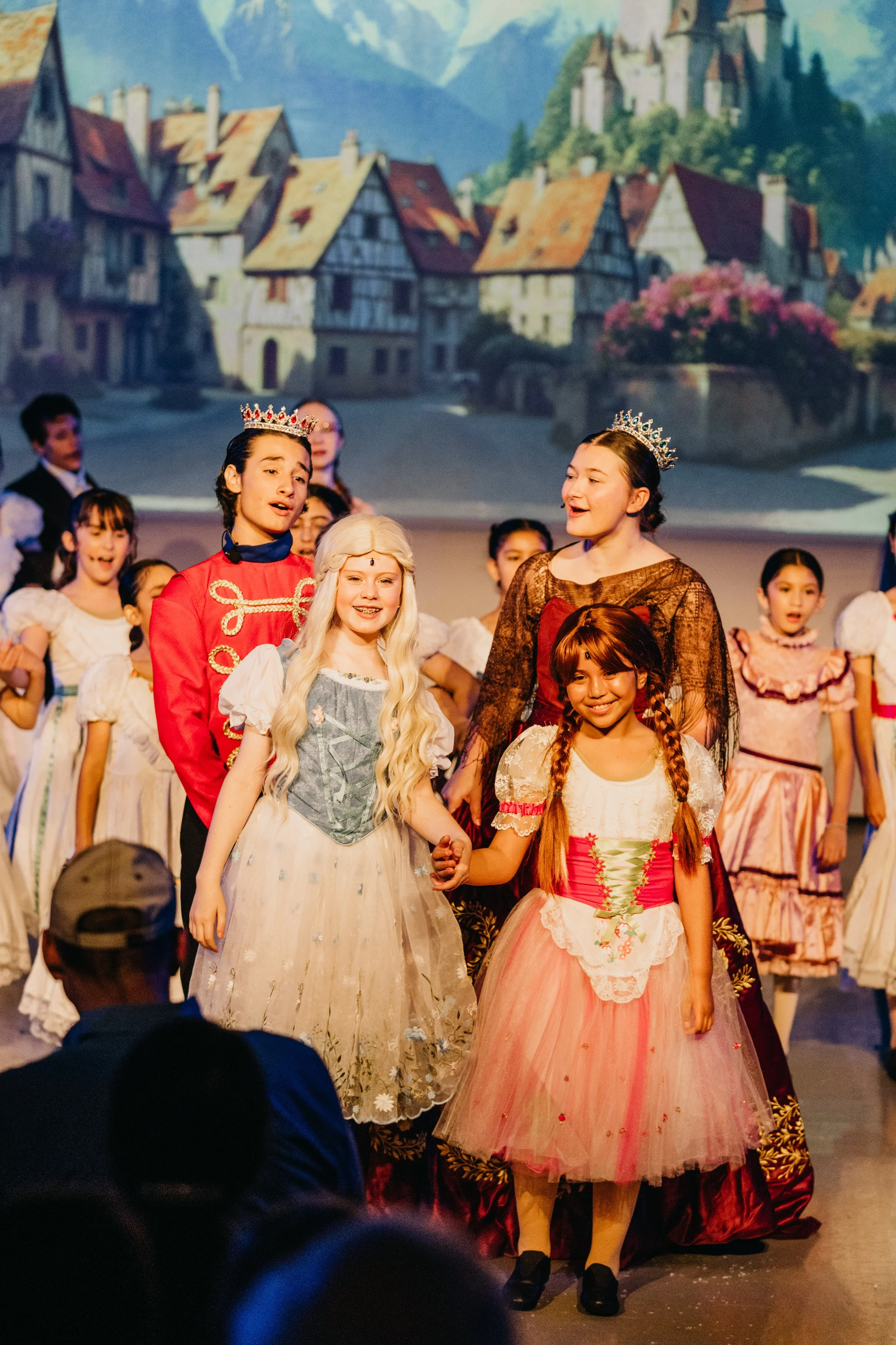 Children dressed in costumes performing on stage with a backdrop of a castle and village scene.