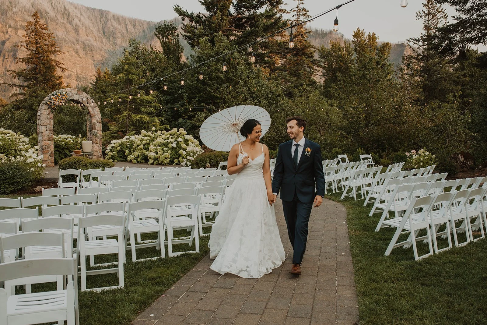 Bride and groom walk hand in hand through an outdoor ceremony space lined with white chairs, string lights, and mountain views at a garden wedding venue.