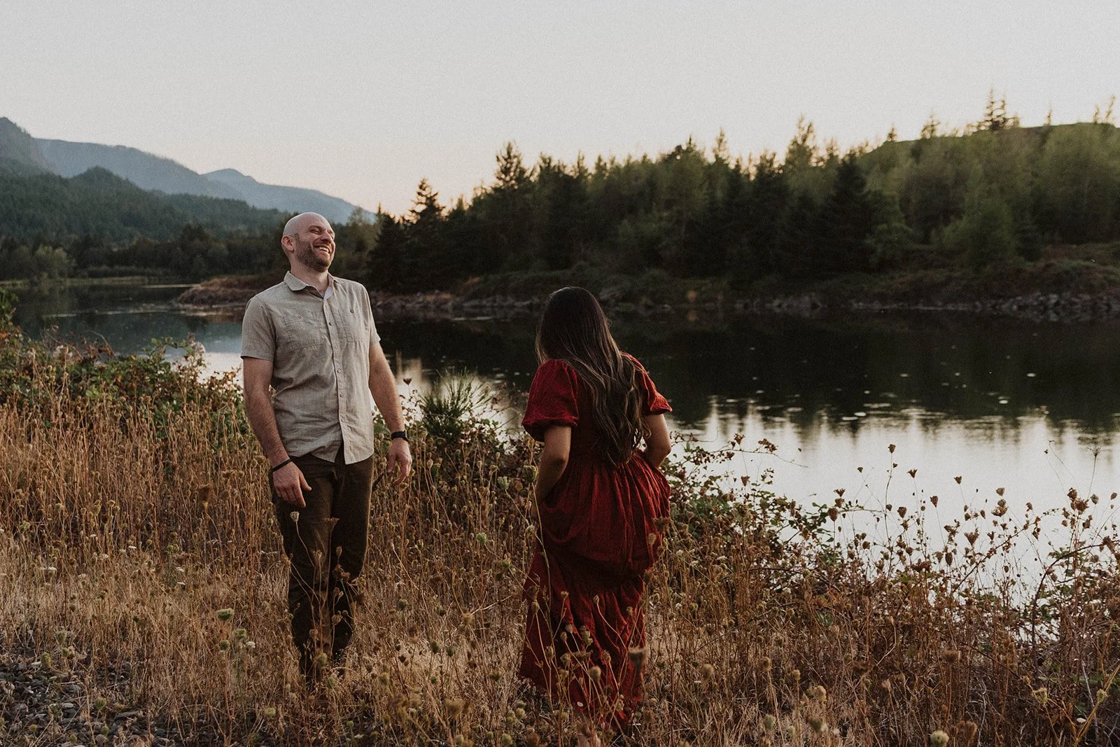 Couple sharing a candid laugh beside a mountain lake during a relaxed outdoor engagement session at sunset.