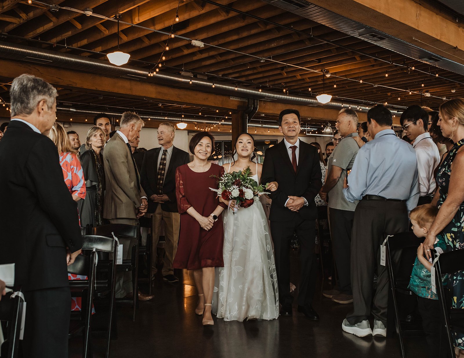 Bride walks down the aisle with her parents during an indoor wedding ceremony surrounded by family and friends in Oregon.