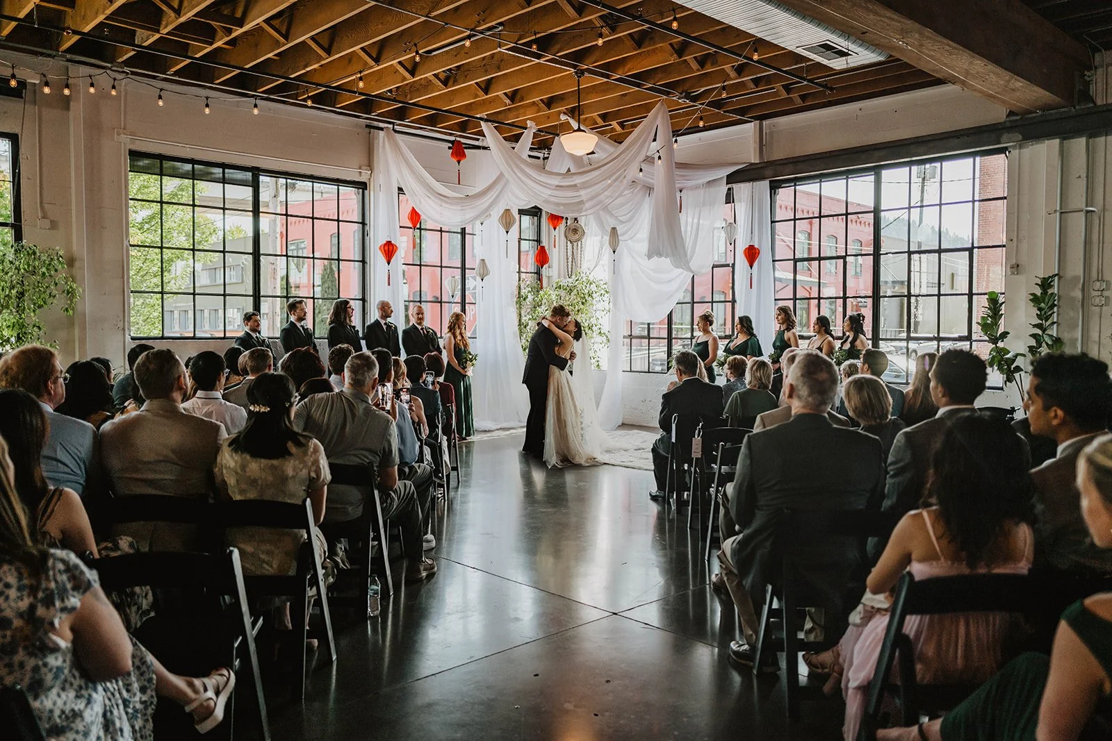 Wide view of wedding guests witnessing a ceremony inside an industrial Portland wedding venue with large windows and draped decor.