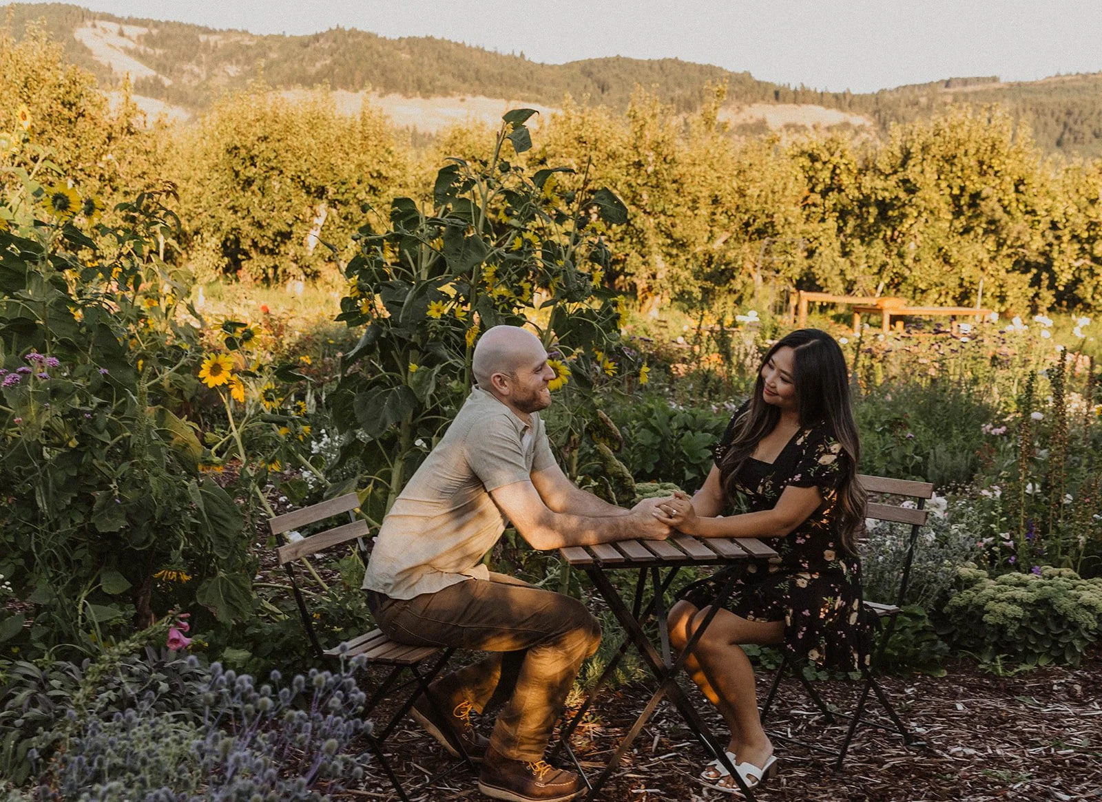 Engaged couple sitting across from each other at a garden table surrounded by flowers and mountain views