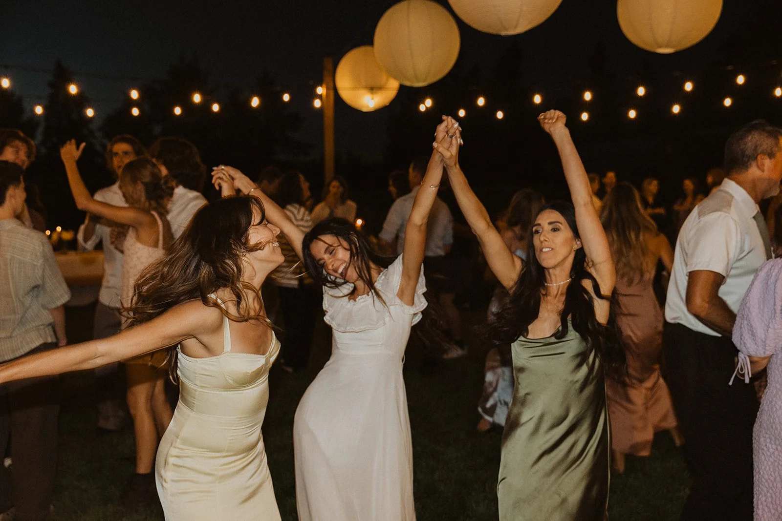 Bride dancing with their arms raised at an outdoor wedding reception at night, surrounded by guests, string lights, and glowing paper lanterns