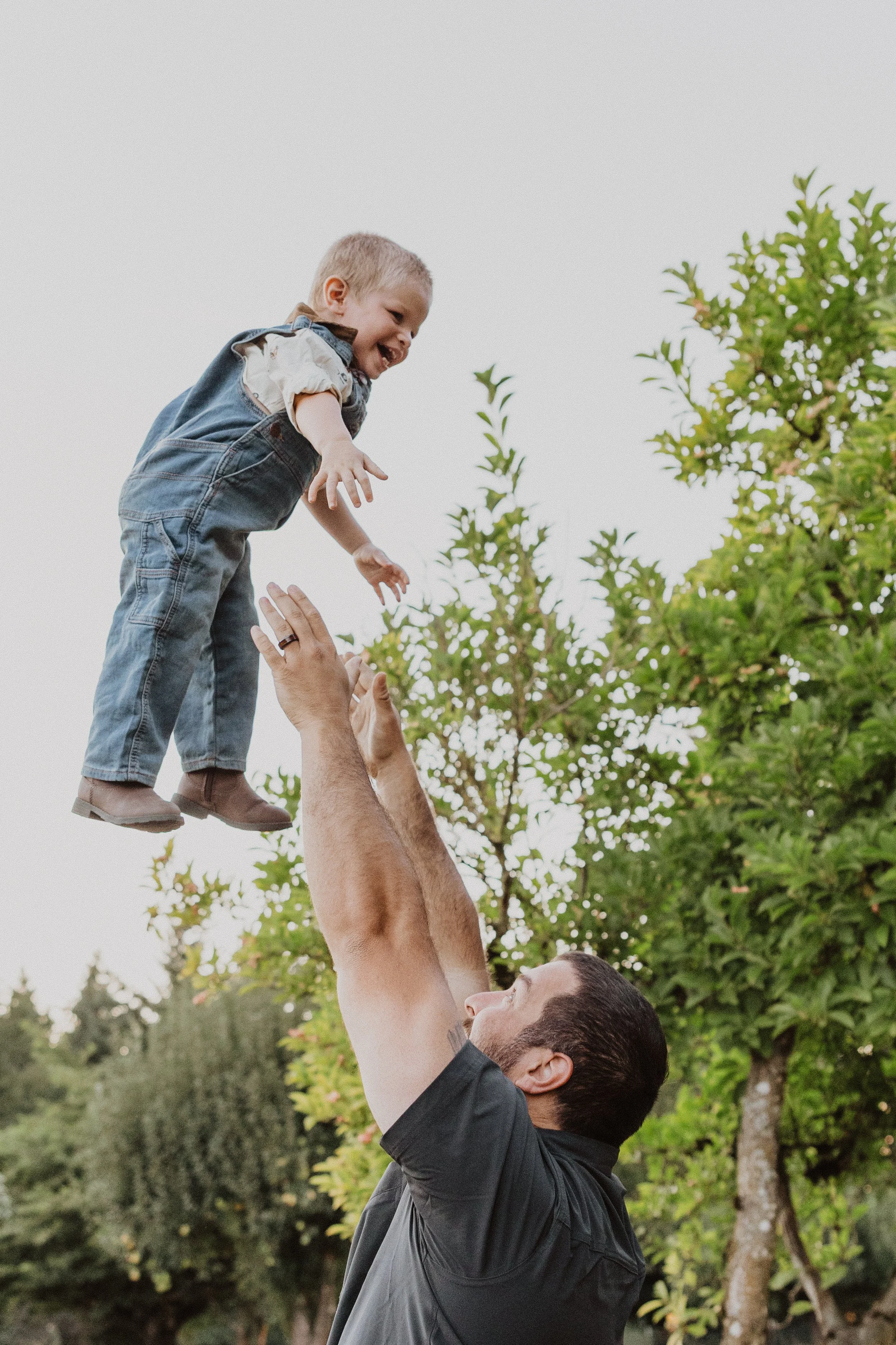 A man lifts a young boy into the air outdoors, surrounded by green trees, with a joyful expression on both their faces.