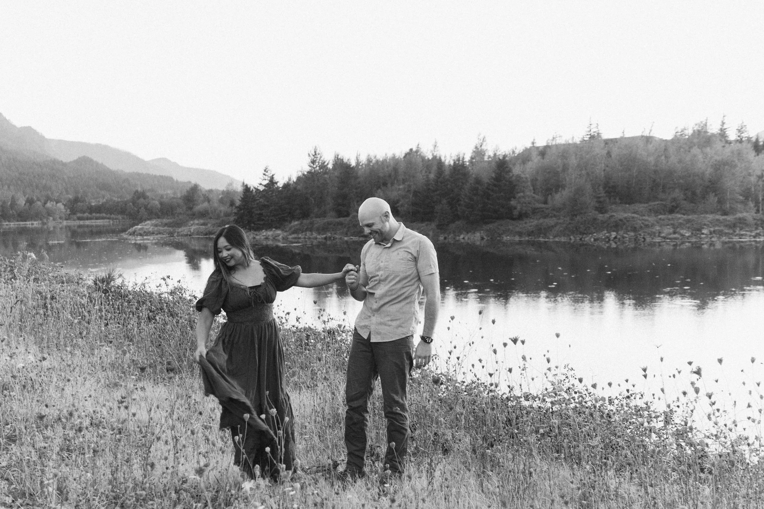 Couple walking hand in hand beside a calm lakeside with mountains in the background, photographed in a documentary black and white style by an Oregon wedding photographer.