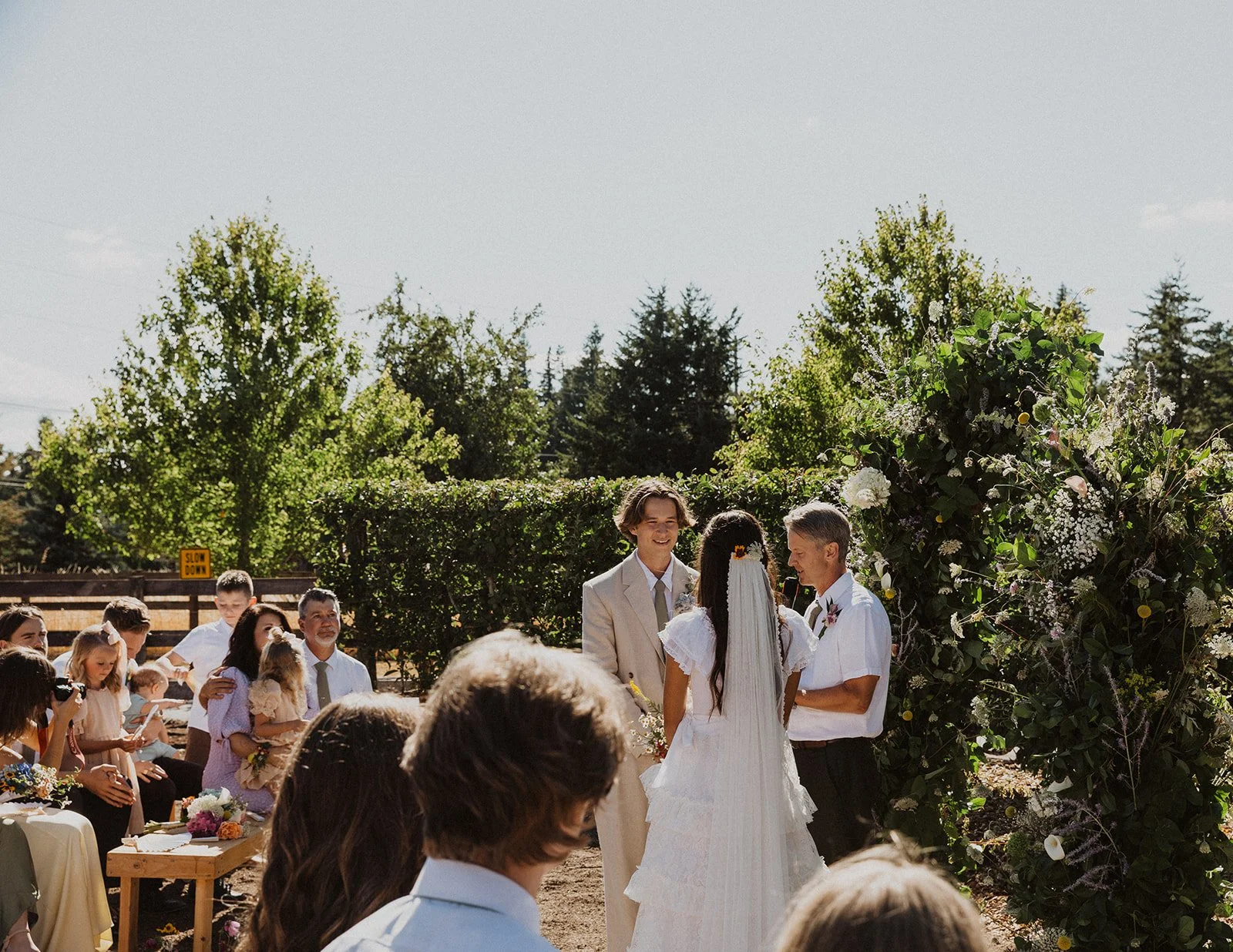 Bride stands facing her seated family and guests during an outdoor ceremony as the officiant speaks, with loved ones watching and holding handheld fans
