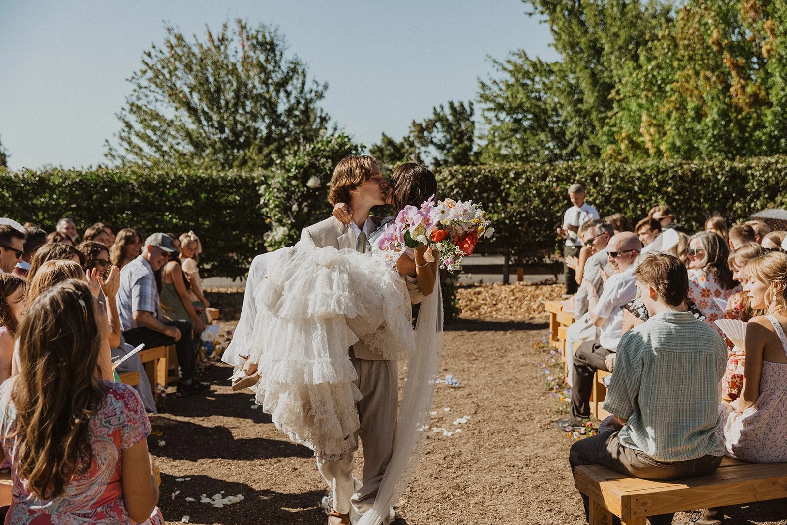 Groom carries and kisses the bride while walking down the aisle at an outdoor wedding, surrounded by smiling guests seated on wooden benches
