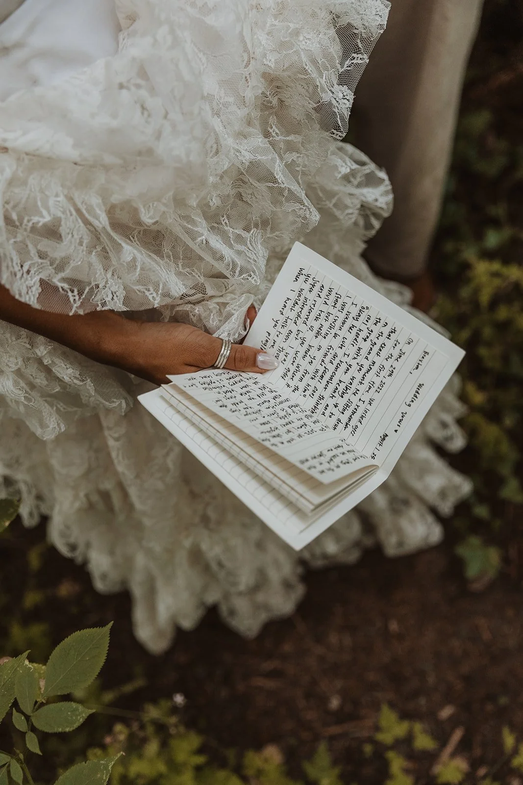 Bride holding handwritten vows in lace dress during outdoor forest wedding ceremony