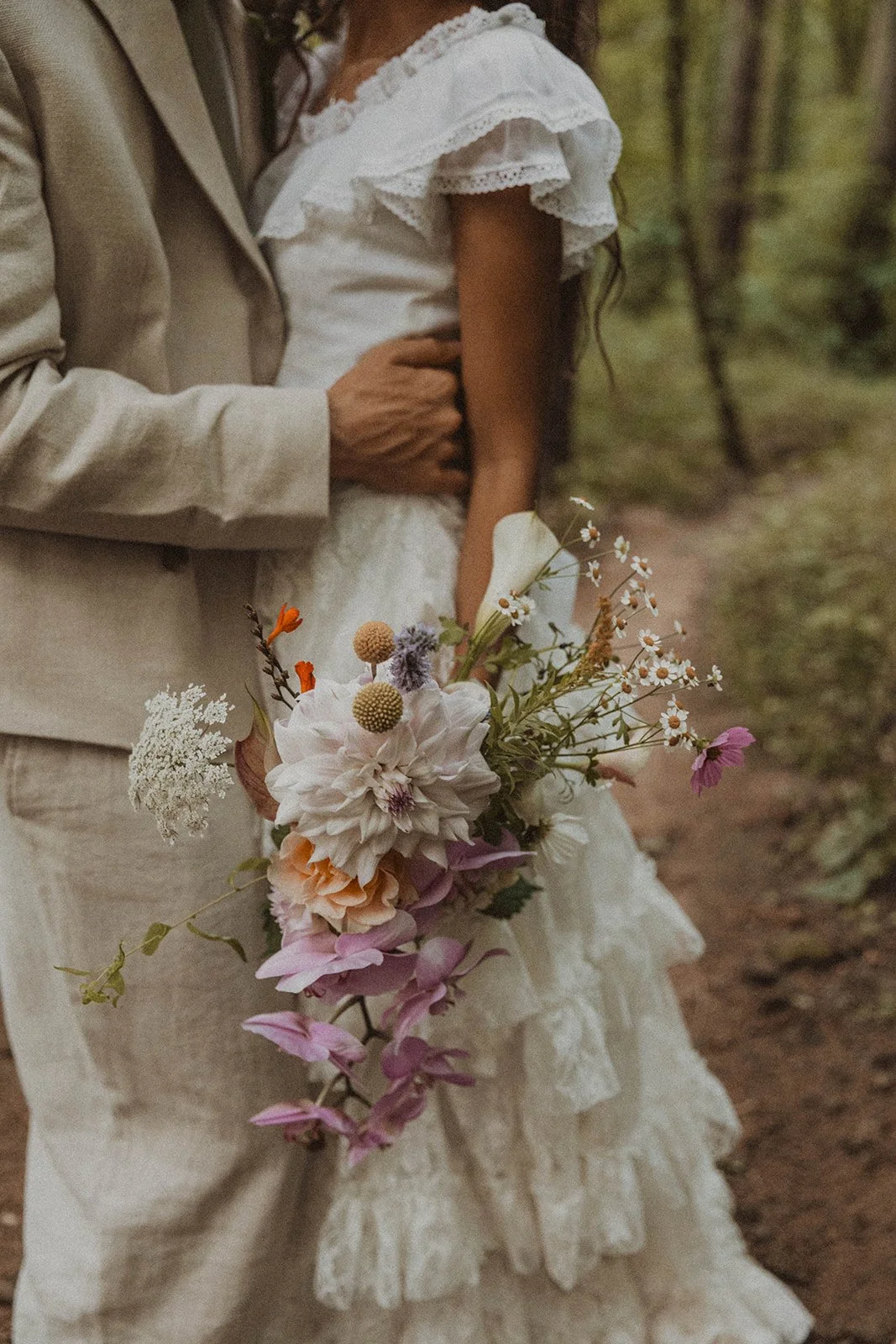 Close-up of bride and groom embracing with colorful wildflower bouquet at forest wedding