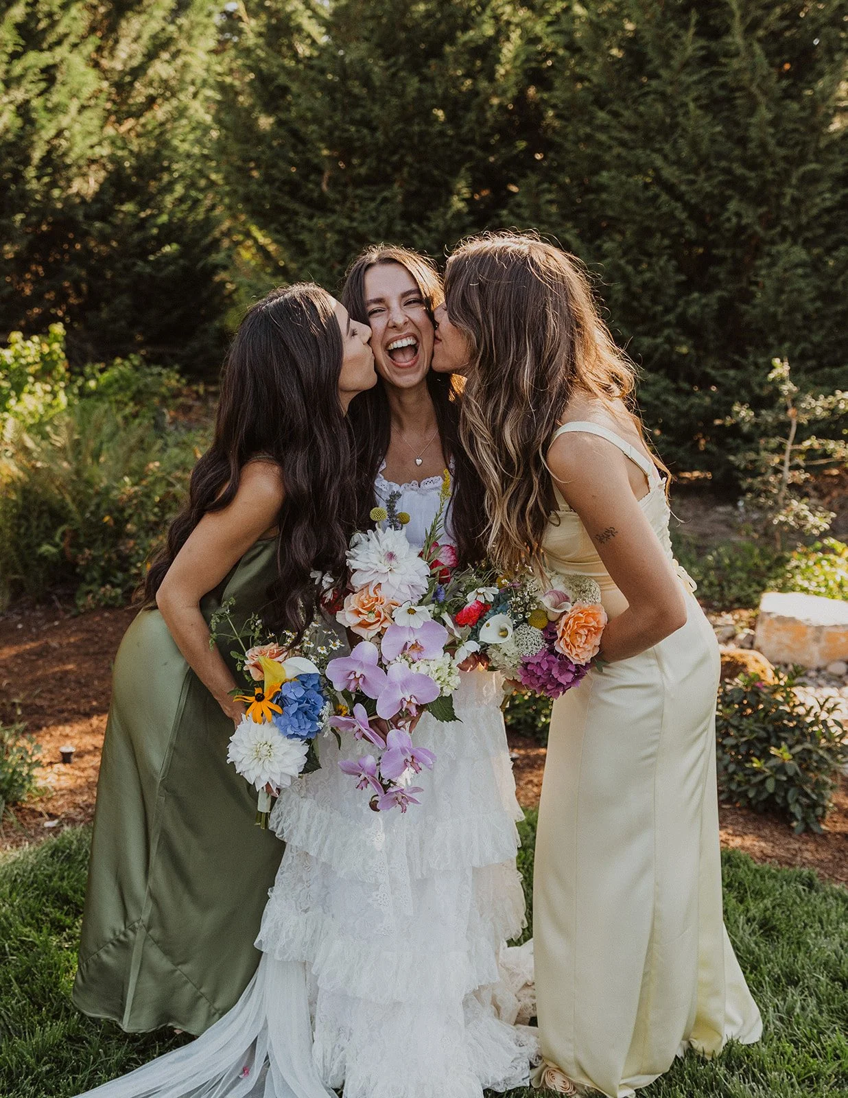 Bride laughing as bridesmaids kiss her cheeks, holding colorful wildflower bouquet outdoors