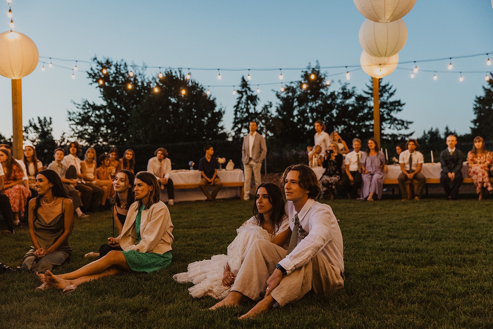 Bride and groom sitting on grass with guests gathered around during outdoor evening wedding reception