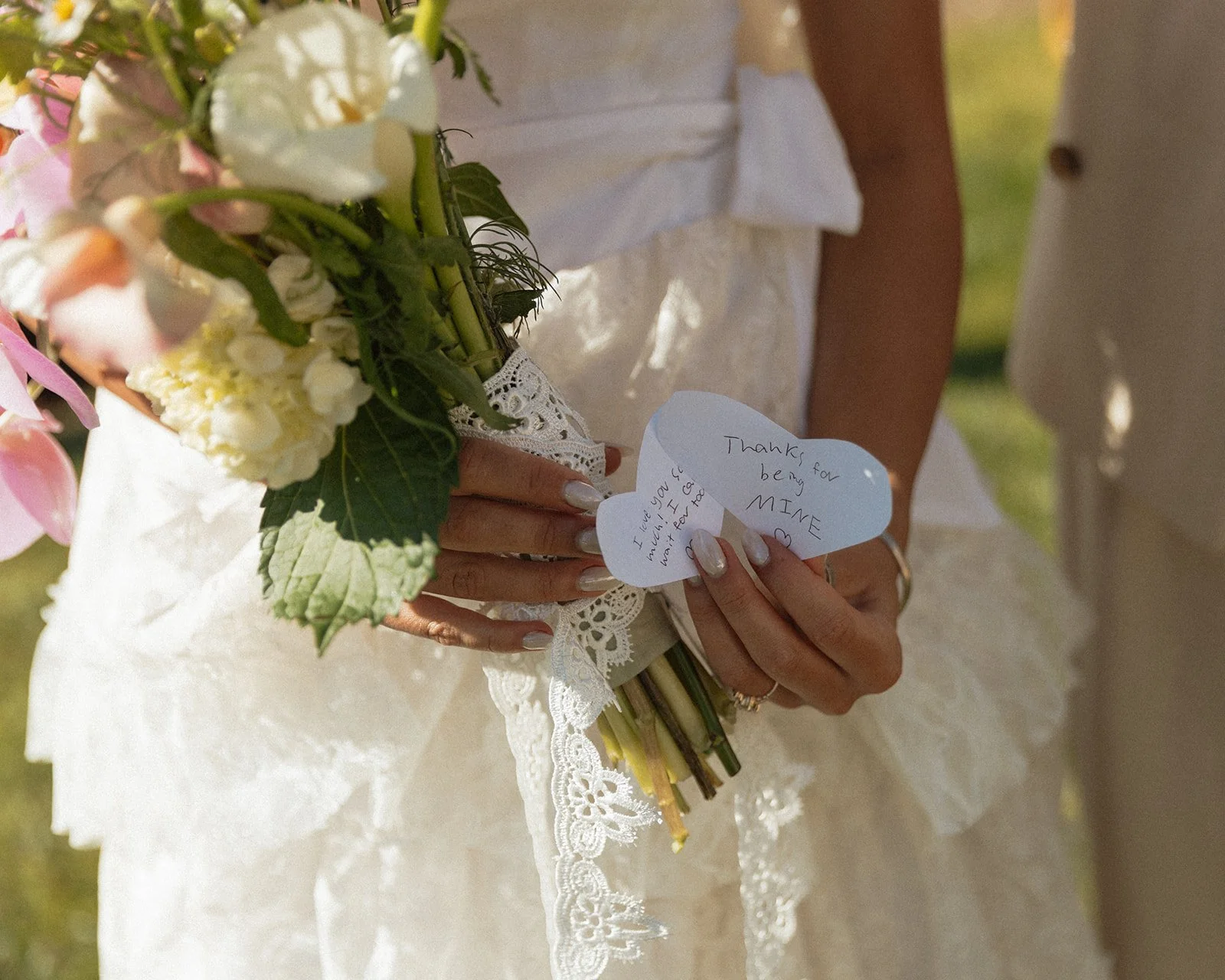 Close-up of bride holding a soft, garden-style bouquet with lace wrap and handwritten wedding vows on heart-shaped paper